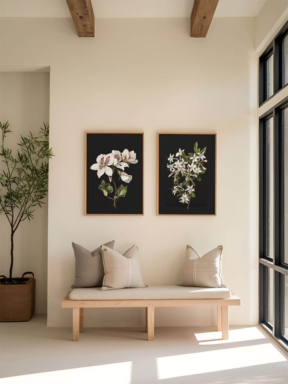 Zwei botanische Kunstdrucke mit weißen Blüten auf schwarzem Hintergrund, symmetrisch an einer weißen Wand aufgehängt. Die Bank darunter ist mit beigen Kissen dekoriert und der Raum wird von natürlichem Licht erhellt, das den minimalistischen Stil unterstreicht.