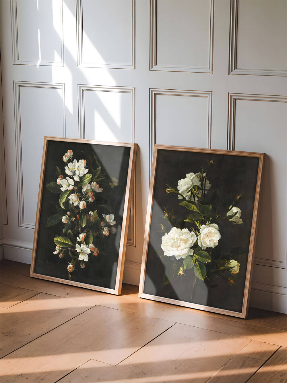 Zwei gerahmte Blumen-Kunstdrucke lehnen in einem hellen Raum an einer getäfelten Wand. Beide Bilder zeigen detaillierte, weiße und rosa Blüten auf einem dunklen, matten Hintergrund. Die Sonnenstrahlen, die durch das Fenster fallen, werfen sanftes Licht auf die Rahmen, wodurch eine warme und behagliche Stimmung erzeugt wird.
