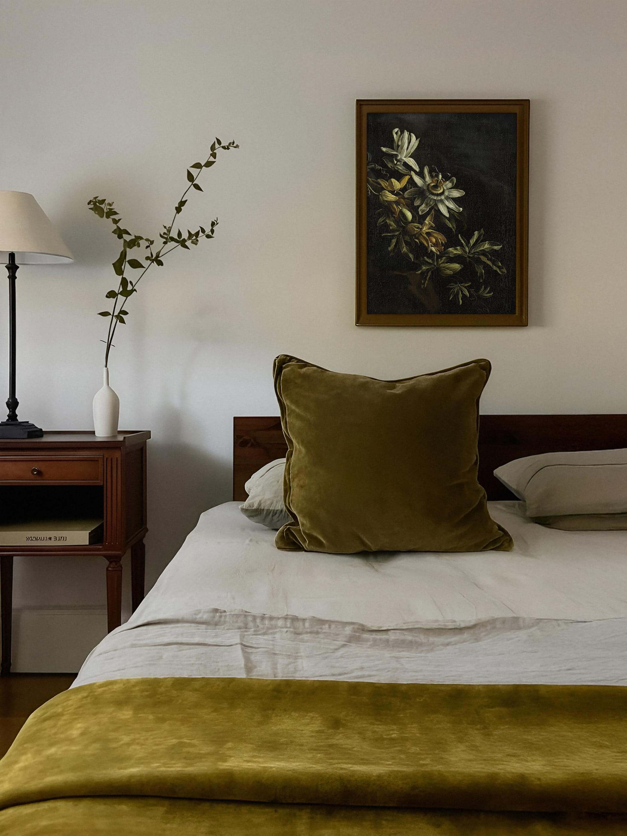 Gerahmter Kunstdruck mit Passionsblume vor schwarzem Hintergrund hängt über einem Bett mit olivgrüner Bettwäsche und Samtkissen. Das dunkle, botanische Motiv fügt sich harmonisch in die warme, gedeckte Schlafzimmergestaltung ein.