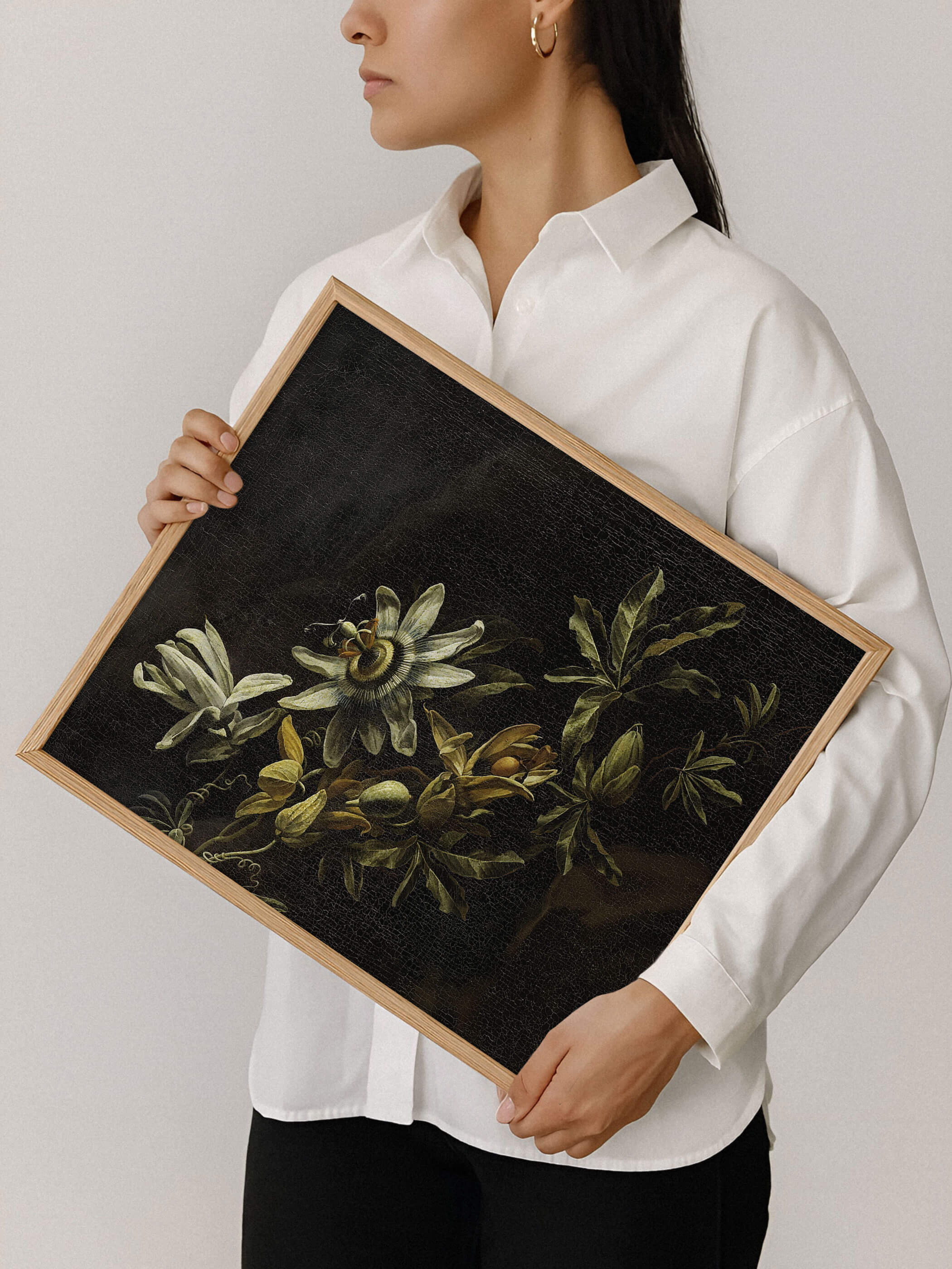 Eine Frau in weißem Hemd hält einen Holzrahmen mit der dunklen Passionsblumen-Malerei vor neutralem Hintergrund. Der Kunstdruck zeigt die feine Textur und den starken Kontrast zwischen heller Blüte und schwarzem Bildgrund im Detail.
