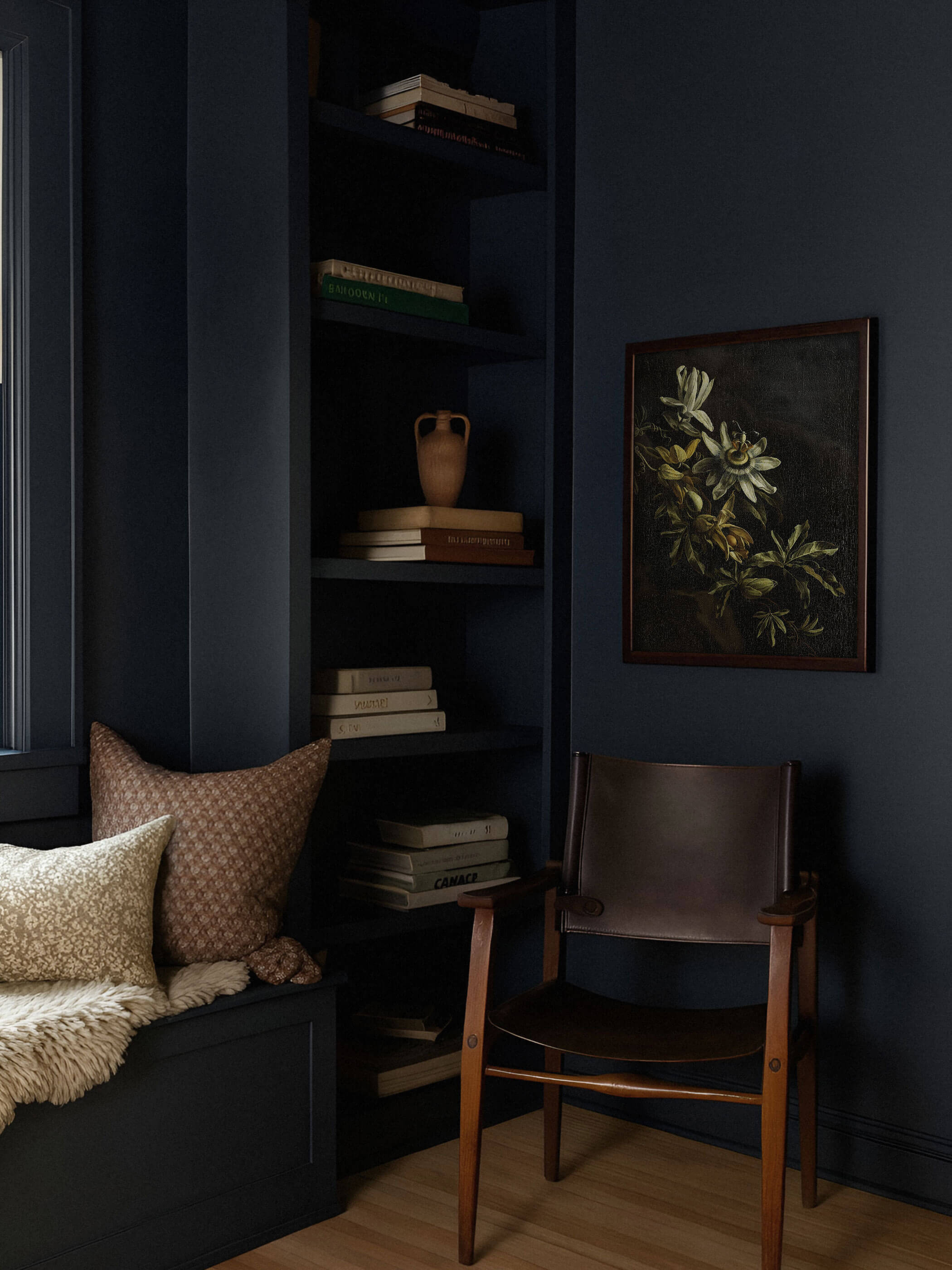 Dunkel gerahmter Kunstdruck mit Passionsblume hängt an einer anthrazitfarbenen Wand neben einem Bücherregal und einem braunen Lederstuhl. Das schwarze Motiv verschmilzt fast mit der Wandfarbe und schafft eine ruhige, moody Leseecke.