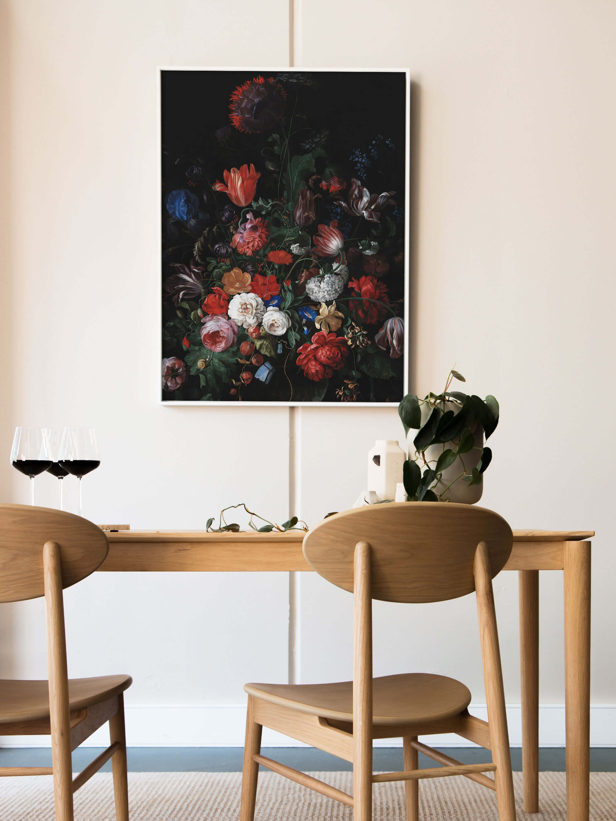 Weiß gerahmter Kunstdruck mit barockem Blumenstillleben lehnt an der Wand über einem Holztisch mit Rotweingläsern und einer grünen Pflanze. Das dunkle, farbintensive Motiv setzt einen stilvollen Akzent im hellen Esszimmer.
