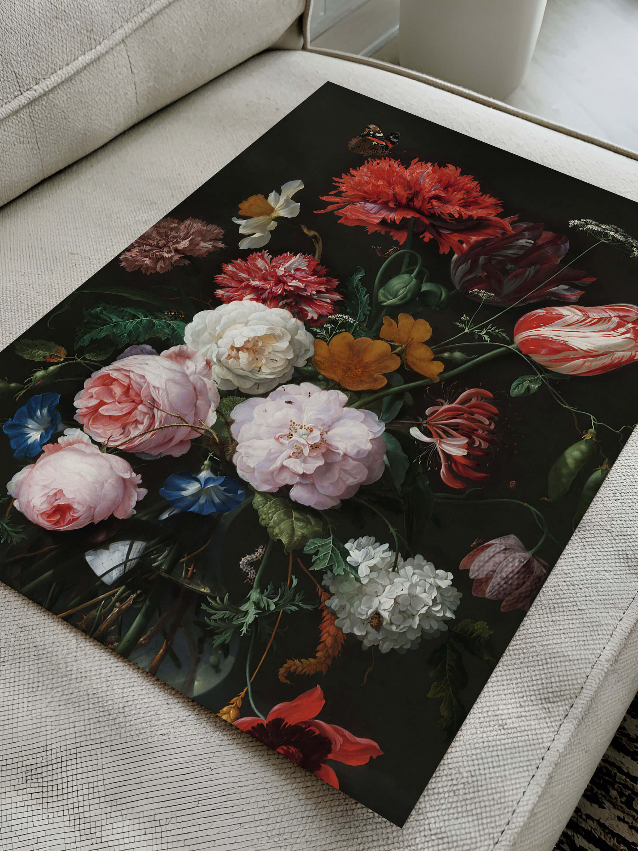 Kunstdruck mit barockem Blumenstillleben aus Mohn, Tulpen und Rosen liegt diagonal auf einem cremefarbenen Sofa. Die Nahaufnahme zeigt die Papierqualität und die kräftigen Farbkontraste des Motivs im Detail.