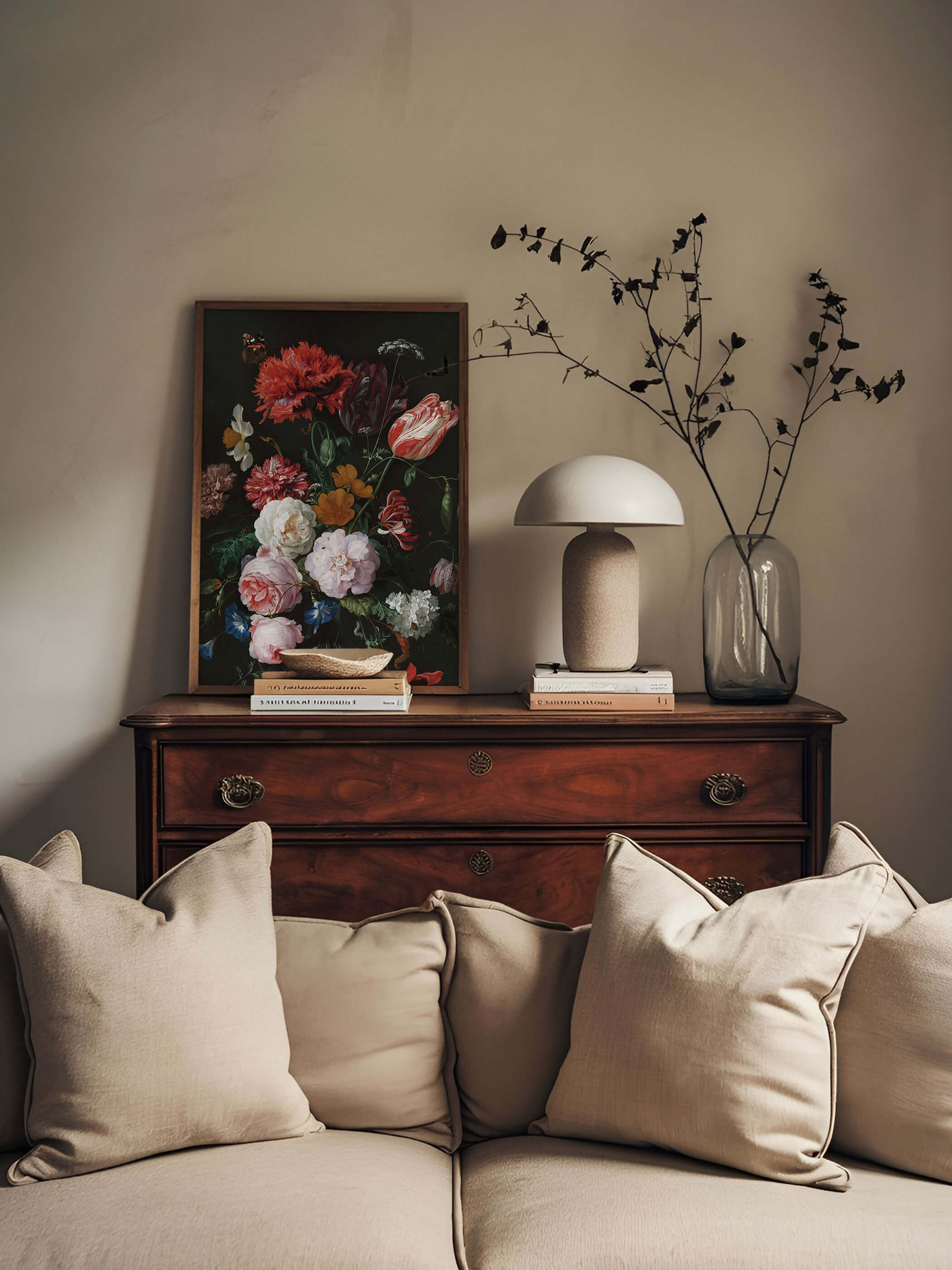 Dunkel gerahmter Kunstdruck mit barockem Blumenstillleben lehnt auf einer antiken Holzkommode neben einer Pilzlampe und einer Glasvase mit Trockenblumen. Das farbenprächtige, dunkle Motiv setzt einen eleganten Akzent hinter dem beigen Sofa.