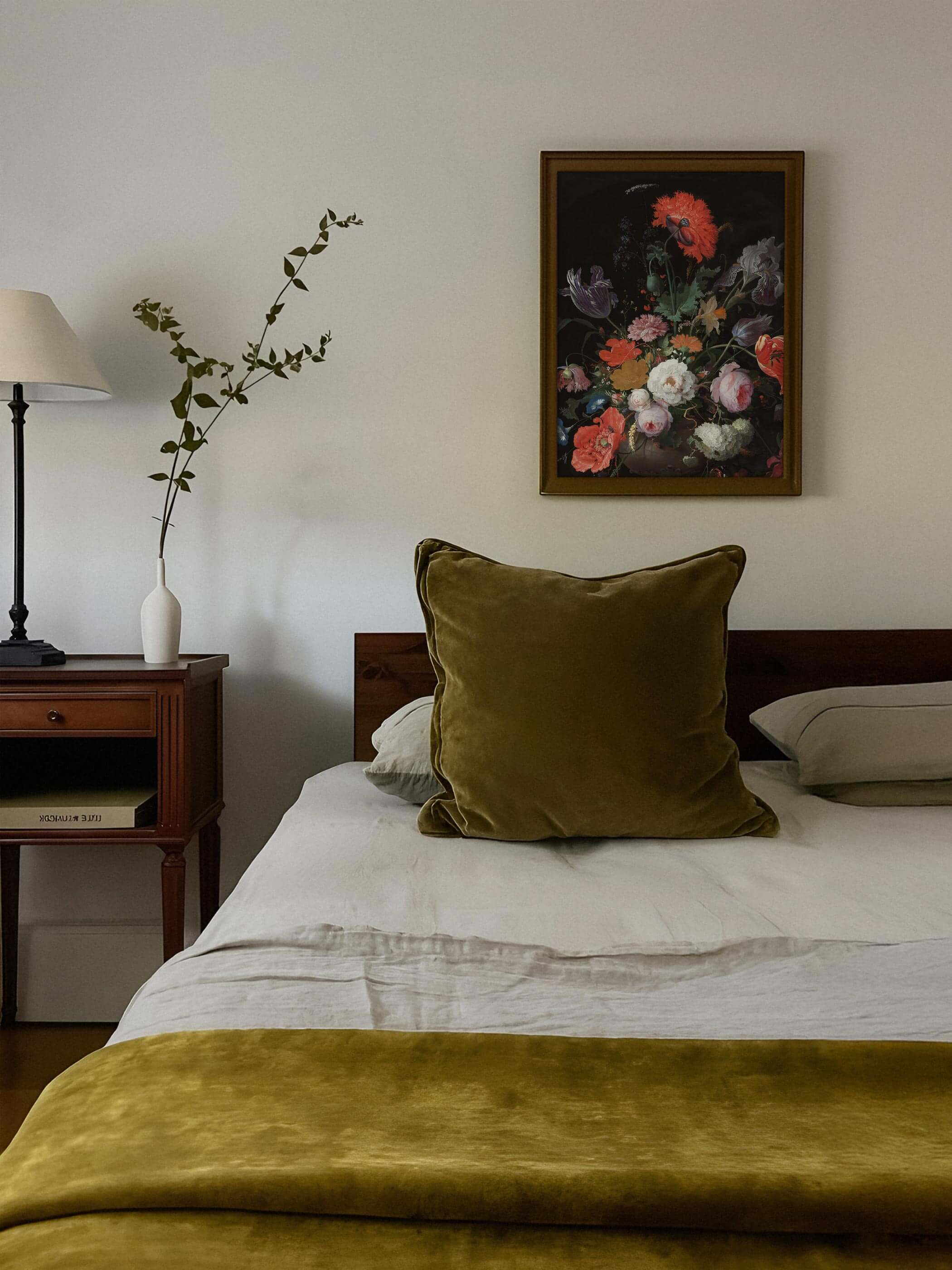 Gerahmter Kunstdruck mit barockem Blumenstillleben hängt über einem Bett mit olivgrünem Samtkissen und weißer Bettwäsche. Das farbenprächtige, dunkle Motiv fügt sich harmonisch in die warme, gedeckte Schlafzimmergestaltung ein.