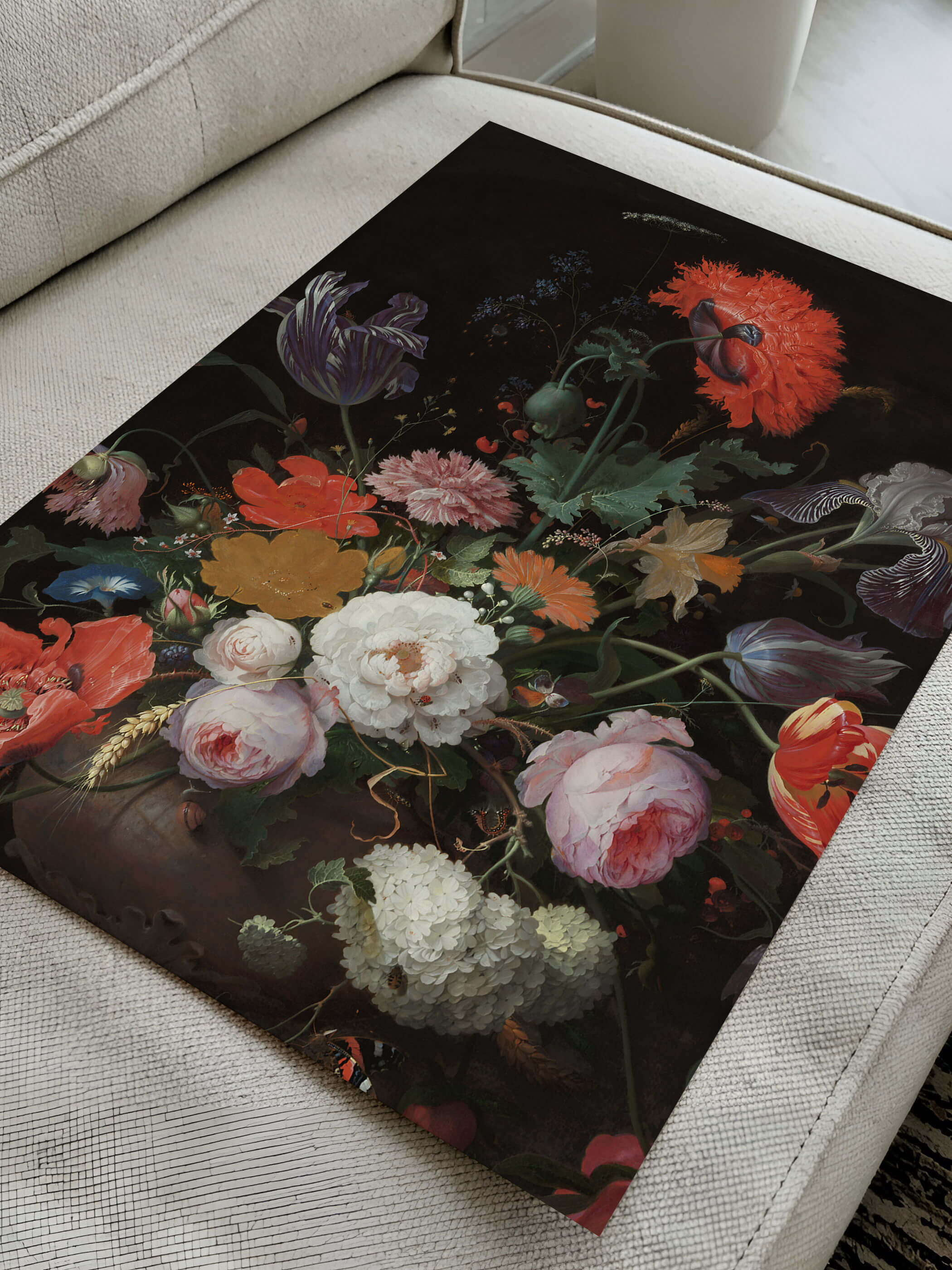 Kunstdruck mit barockem Blumenstillleben aus Mohn, Iris und Pfingstrosen liegt diagonal auf einem cremefarbenen Sofa. Die Nahaufnahme zeigt die Papierqualität und die kräftigen Farbkontraste des Motivs im Detail.