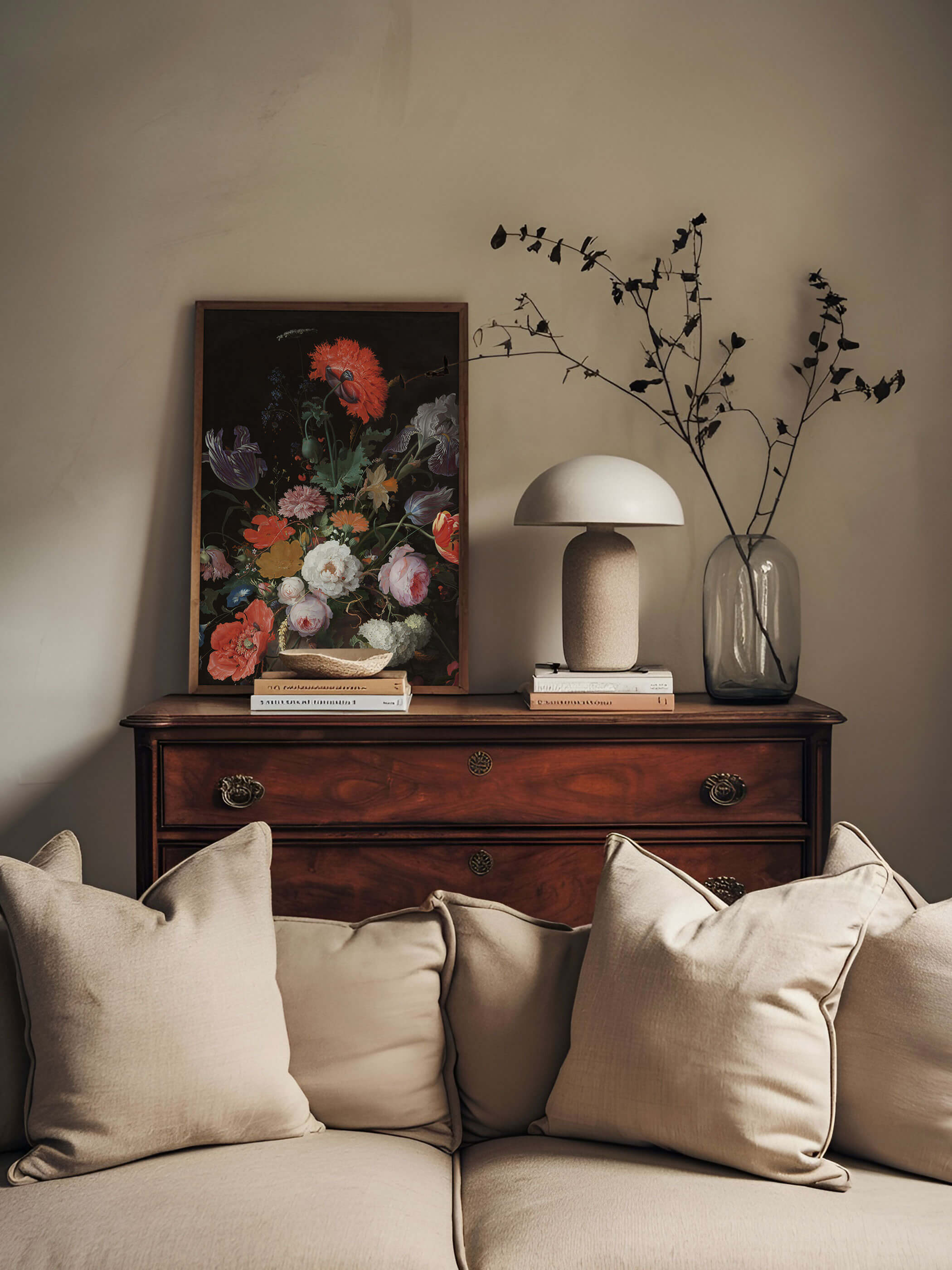 Dunkel gerahmter Kunstdruck mit barockem Blumenstillleben lehnt auf einer antiken Holzkommode neben einer Pilzlampe und einer Glasvase mit Trockenblumen. Das farbenprächtige, dunkle Motiv setzt einen eleganten Akzent hinter dem beigen Sofa.