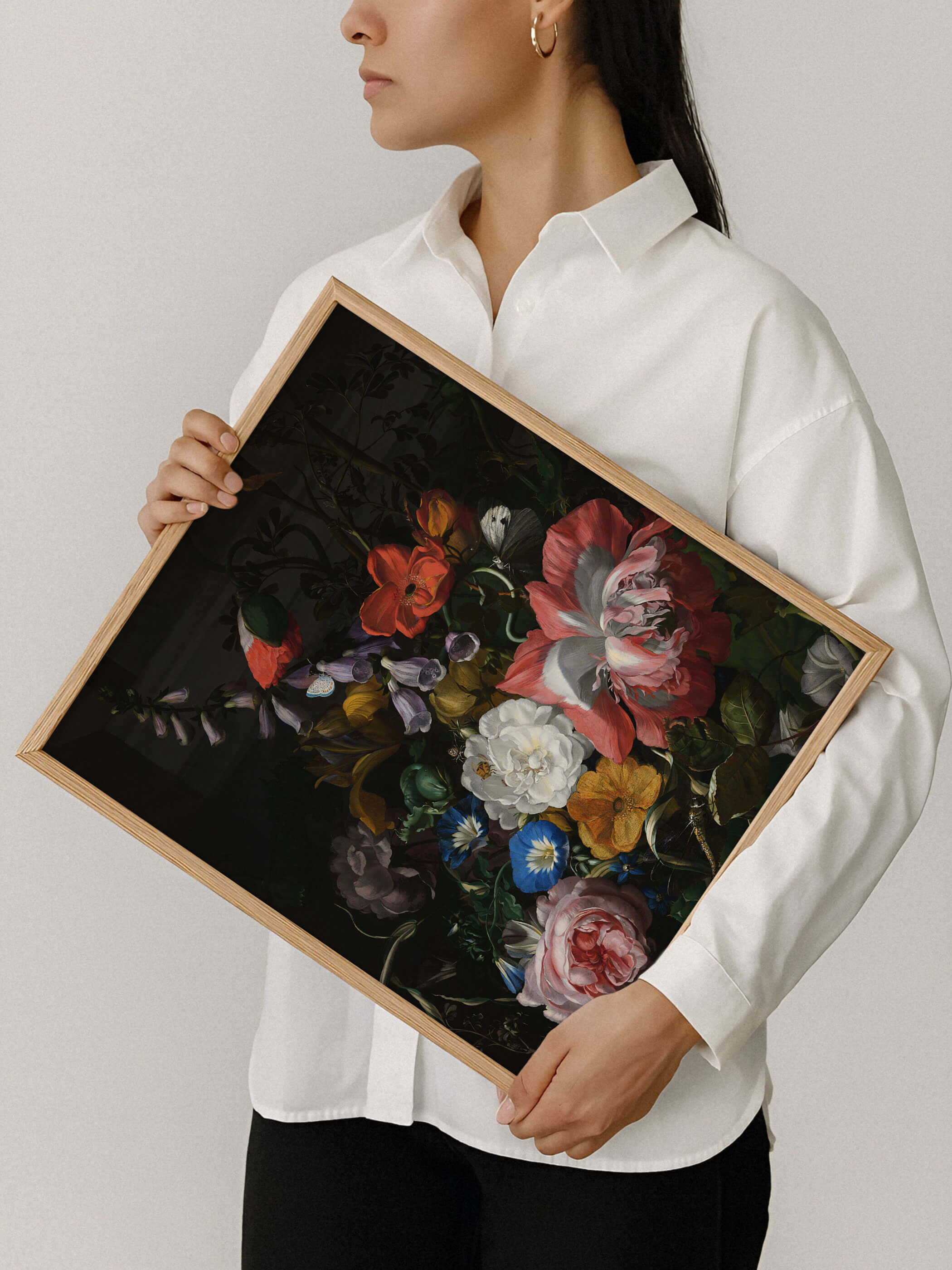 Eine Frau in weißem Hemd hält einen Holzrahmen mit dem barocken Blumenstillleben aus Rosen, Mohn und Prunkwinde vor neutralem Hintergrund. Der Kunstdruck zeigt die kräftigen Farben und die feine Rahmung im Detail.