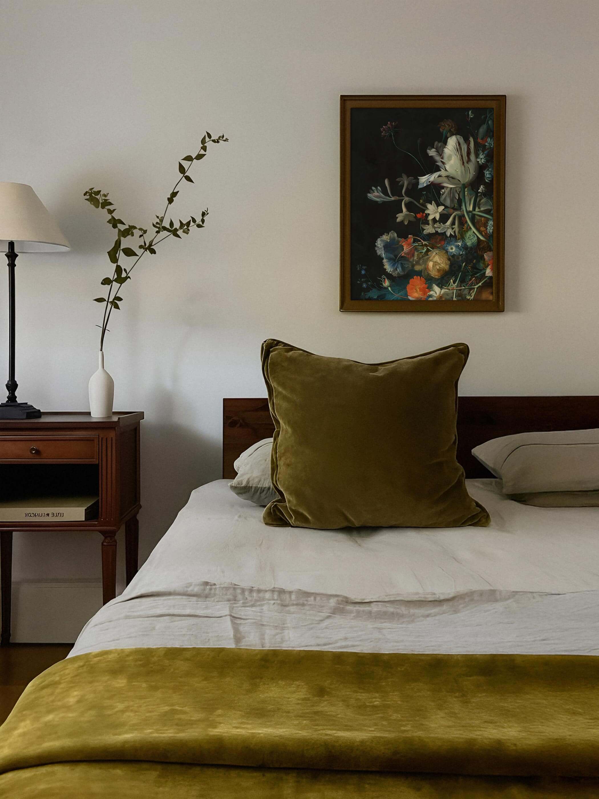 Golden gerahmter Kunstdruck mit barockem Blumenstillleben hängt über einem Bett mit olivgrünem Samtkissen und weißer Bettwäsche. Das dunkle, ruhige Motiv passt zur warmen, gedeckten Schlafzimmerfarbgebung.