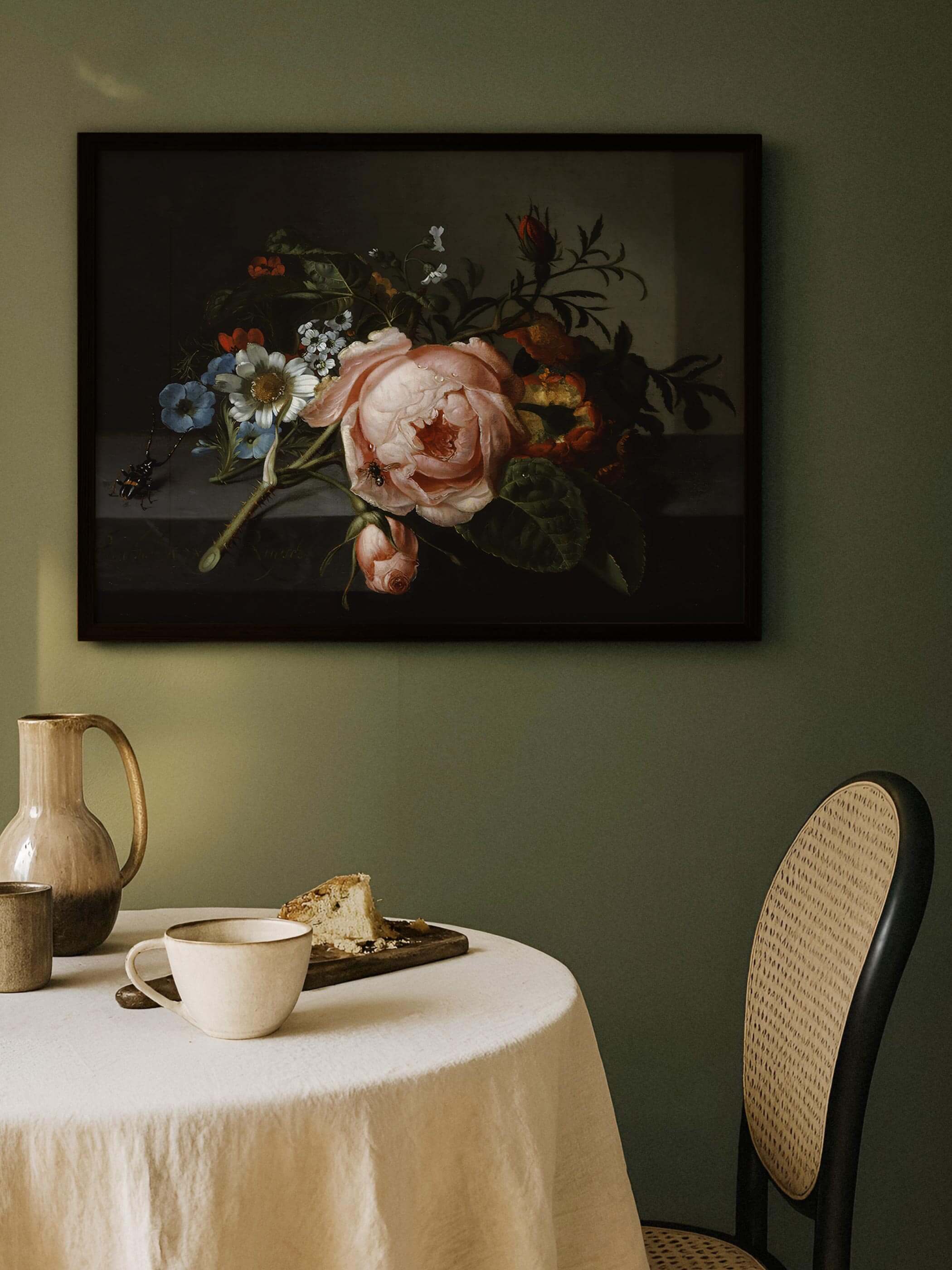 Dunkel gerahmter Kunstdruck mit Rosenzweig hängt an einer olivgrünen Wand über einem gedeckten Tisch mit Kaffeetasse und Kuchen. Das ruhige, reduzierte Motiv setzt einen stimmungsvollen Akzent im gemütlichen Esszimmer.
