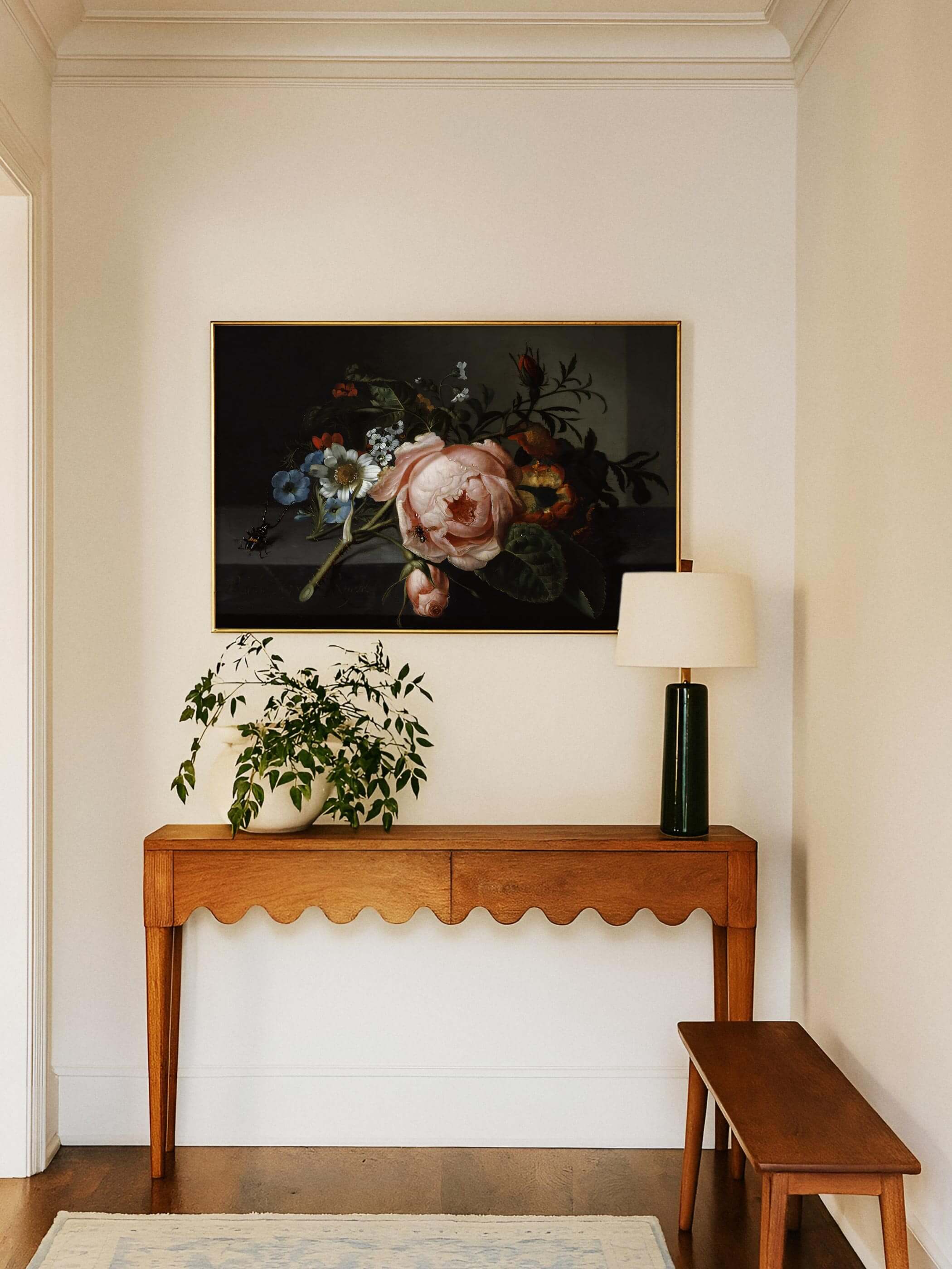 Golden gerahmter Kunstdruck mit Rosenzweig und Käfer hängt über einer Holzkonsole mit Pflanze und Tischlampe im hellen Flur. Das dunkle, ruhige Motiv setzt einen eleganten Akzent neben der Bank und dem weichen Teppich.