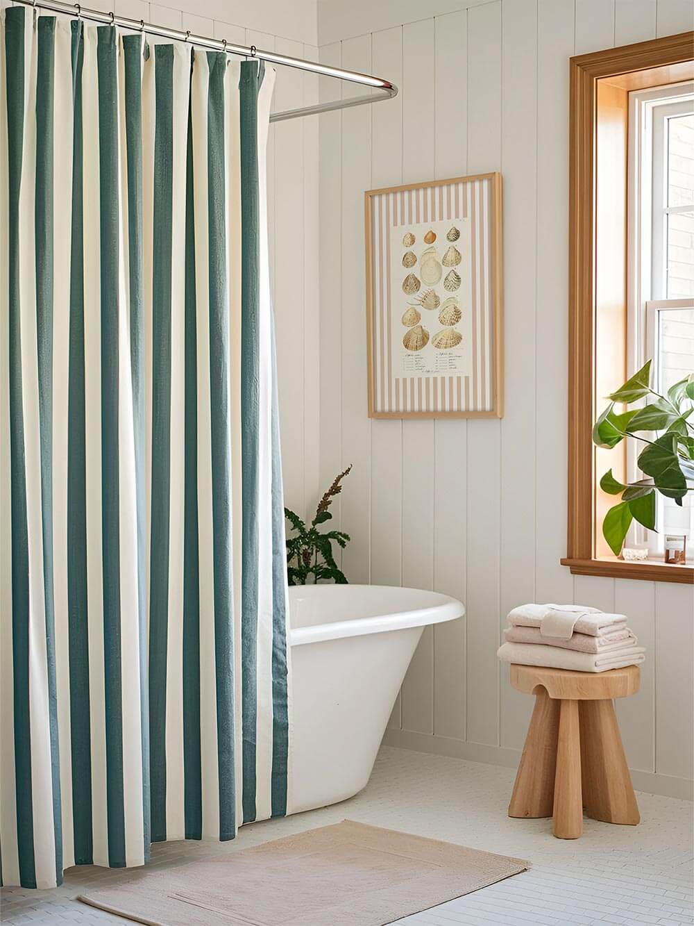 Vintage-Muschel-Kunstdruck mit zarten Naturfarben und roséfarbenem Streifenrahmen an weißer Badezimmerwand. Maritime Wanddeko neben einer freistehenden Badewanne mit gestreiftem Duschvorhang.

