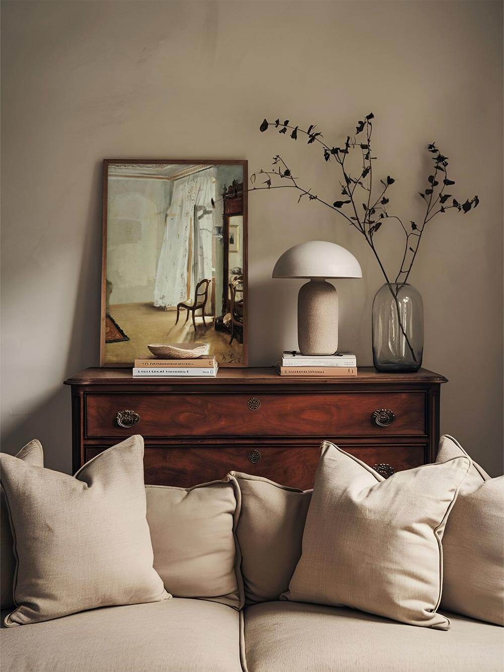 Vintage-Interieur-Kunstdruck auf einer Kommode dekoriert mit Büchern, Keramikvasen und Trockenblumen. Die nostalgische Darstellung eines lichtdurchfluteten Zimmers bringt klassische Eleganz und eine beruhigende Atmosphäre in den Raum.