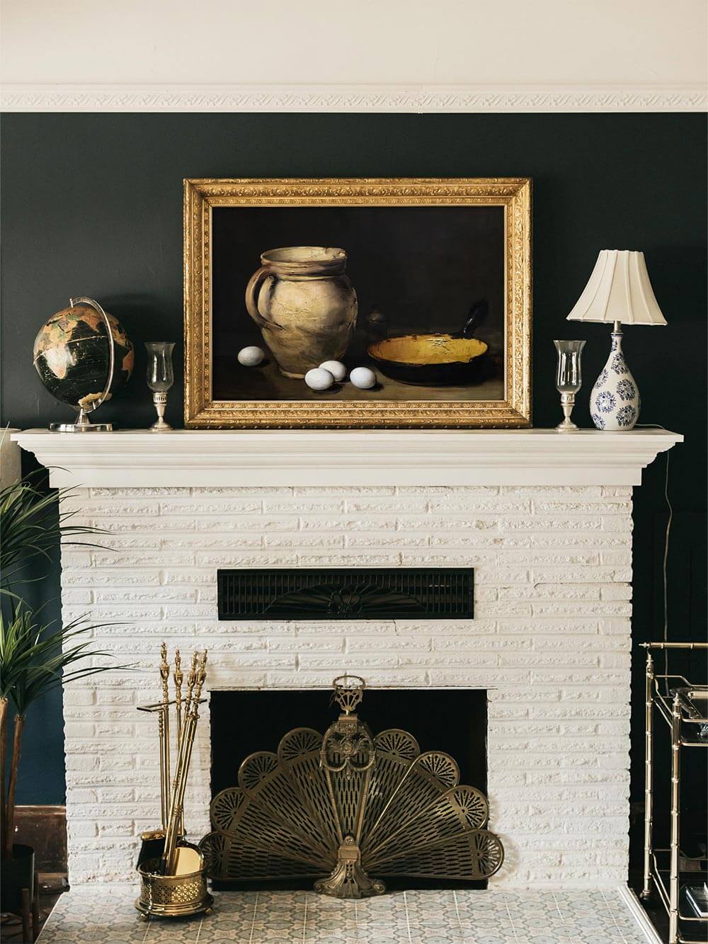 Kunstdruck eines rustikalen Stilllebens mit Keramikkrug, Eiern und Pfanne, eingerahmt in Gold, über einem Kamin auf dunkelgrüner Wand – klassische Wandkunst für ein elegantes Vintage-Interieur.