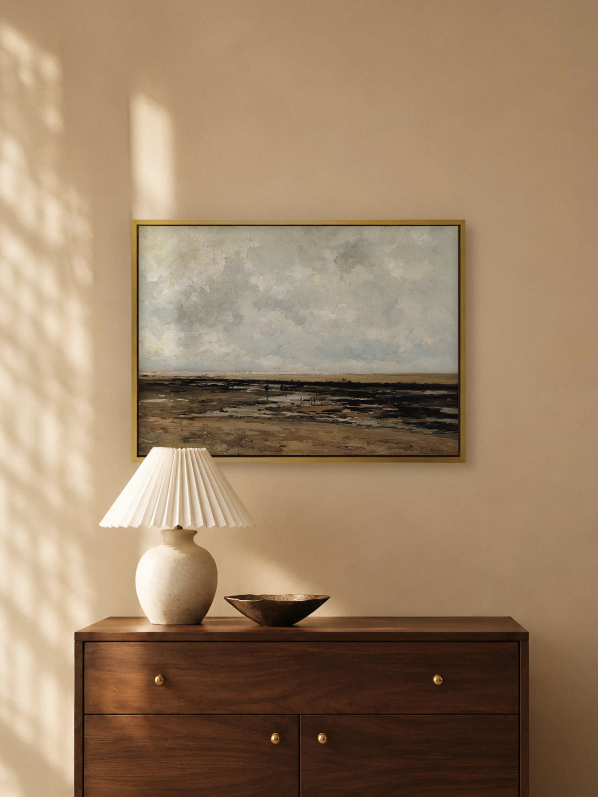 Maritimes Leinwandbild im goldenen Rahmen über einer dunklen Holzkommode mit Messingknöpfen. Auf der Kommode stehen eine Keramiklampe mit weißem Plisseeschirm und eine flache Bronzeschale, warmes Licht fällt auf die Wand.