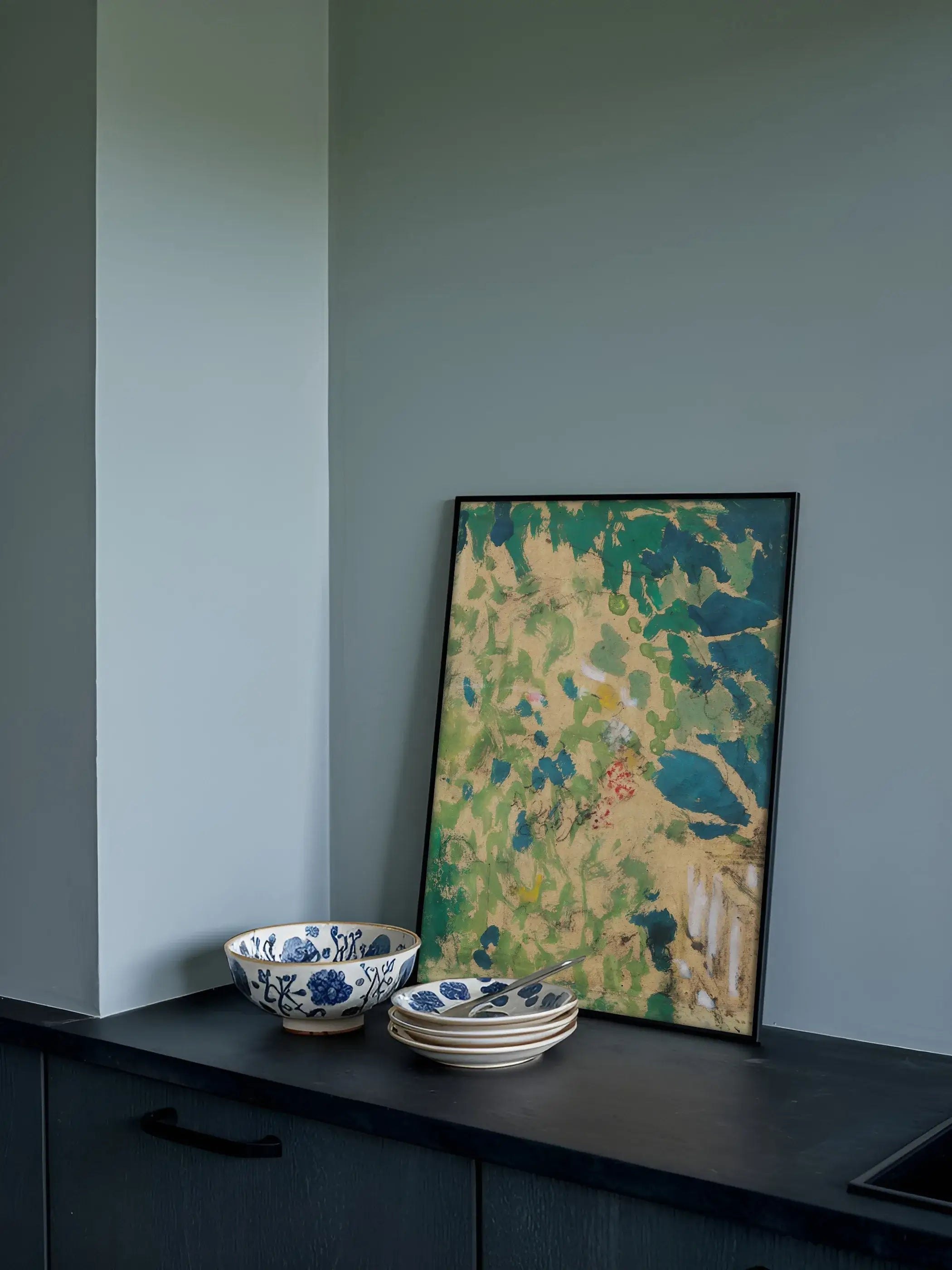 Wandbild mit grün-blauer abstrakter Landschaft lehnt gerahmt an einer dunkelblauen Küchenwand neben blau-weiß gemustertem Geschirr. Das lebendige Wandbild passt zu einem warmen, naturnahen Küchenstil.