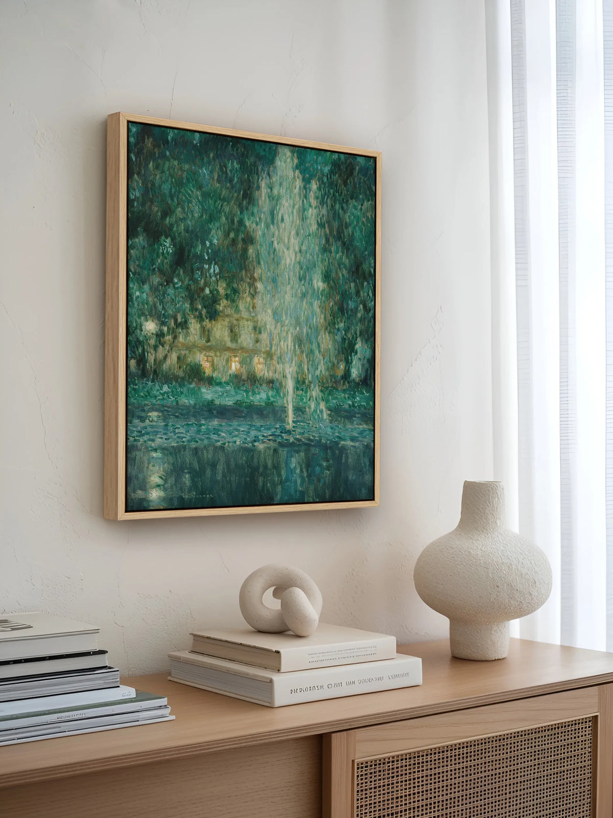 Grünes Landschaft Leinwandbild im Naturholzrahmen über einem hellen Sideboard mit Rattanfront. Davor stehen eine strukturierte Keramikvase, eine Knotenskulptur und gestapelte Bücher, daneben fällt Licht durch einen weißen Vorhang.