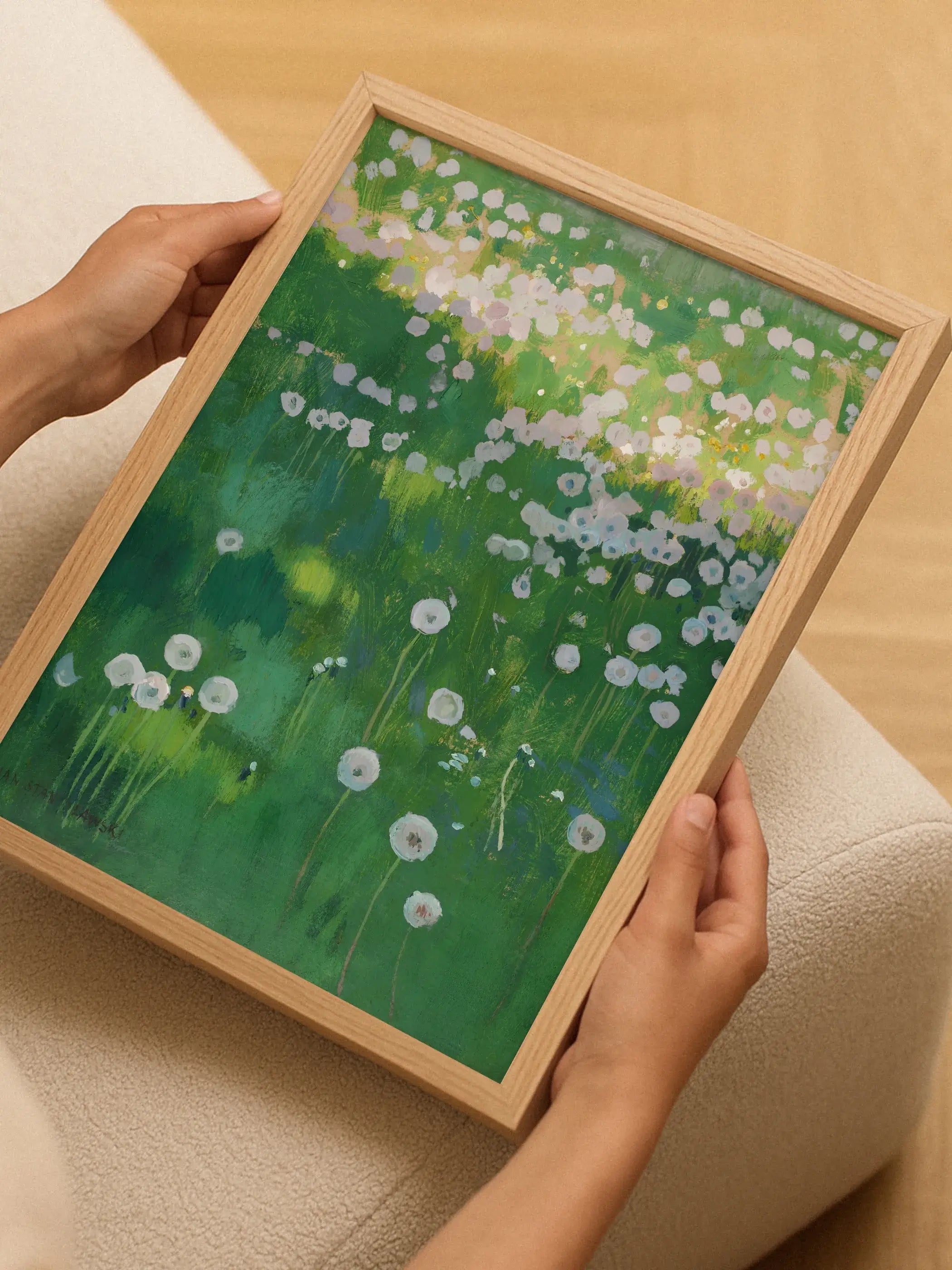 Gerahmter Landschafts-Kunstdruck mit Löwenzahnwiese wird von zwei Händen gehalten, im Hintergrund helles Holz und ein beigefarbenes Polstermöbel. Das frische, grüne Wandbild wirkt einladend und naturverbunden.