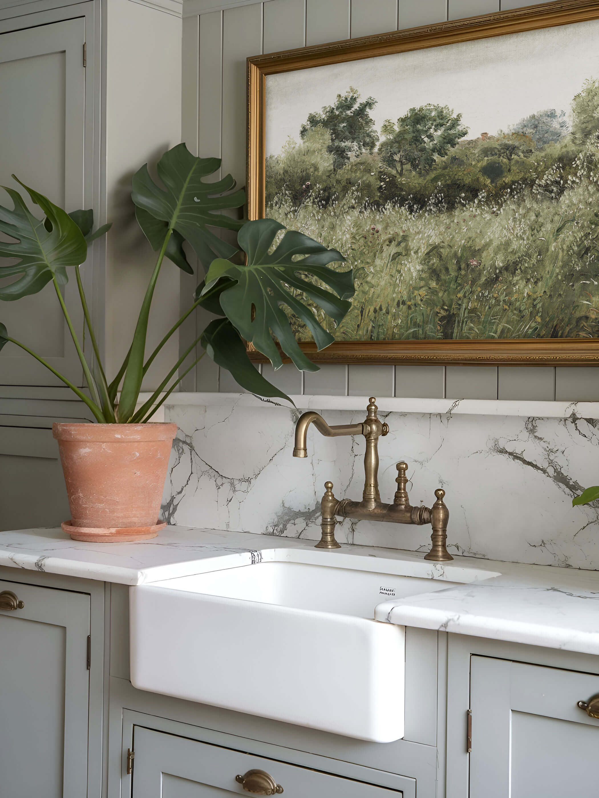 Vintage Landschaftsdruck in goldgerahmtem Bild über weißer Marmorarbeitsplatte in einer Landhausküche mit Messingarmaturen und einer Monstera-Pflanze.