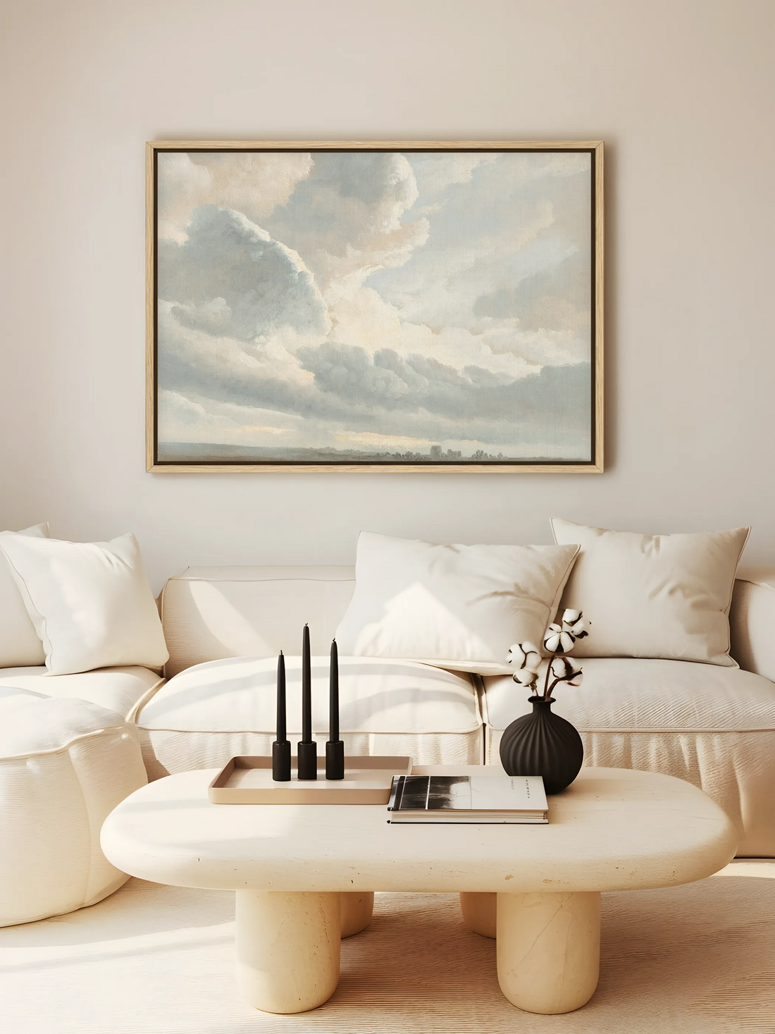 Die Vintage Wolkenlandschaft hängt als Leinwandbild im Naturholzrahmen über einem cremefarbenen Sofa mit großen Kissen. Auf dem rundlichen Couchtisch aus hellem Stein stehen drei schwarze Kerzen und eine schwarze Vase mit Baumwollzweigen.