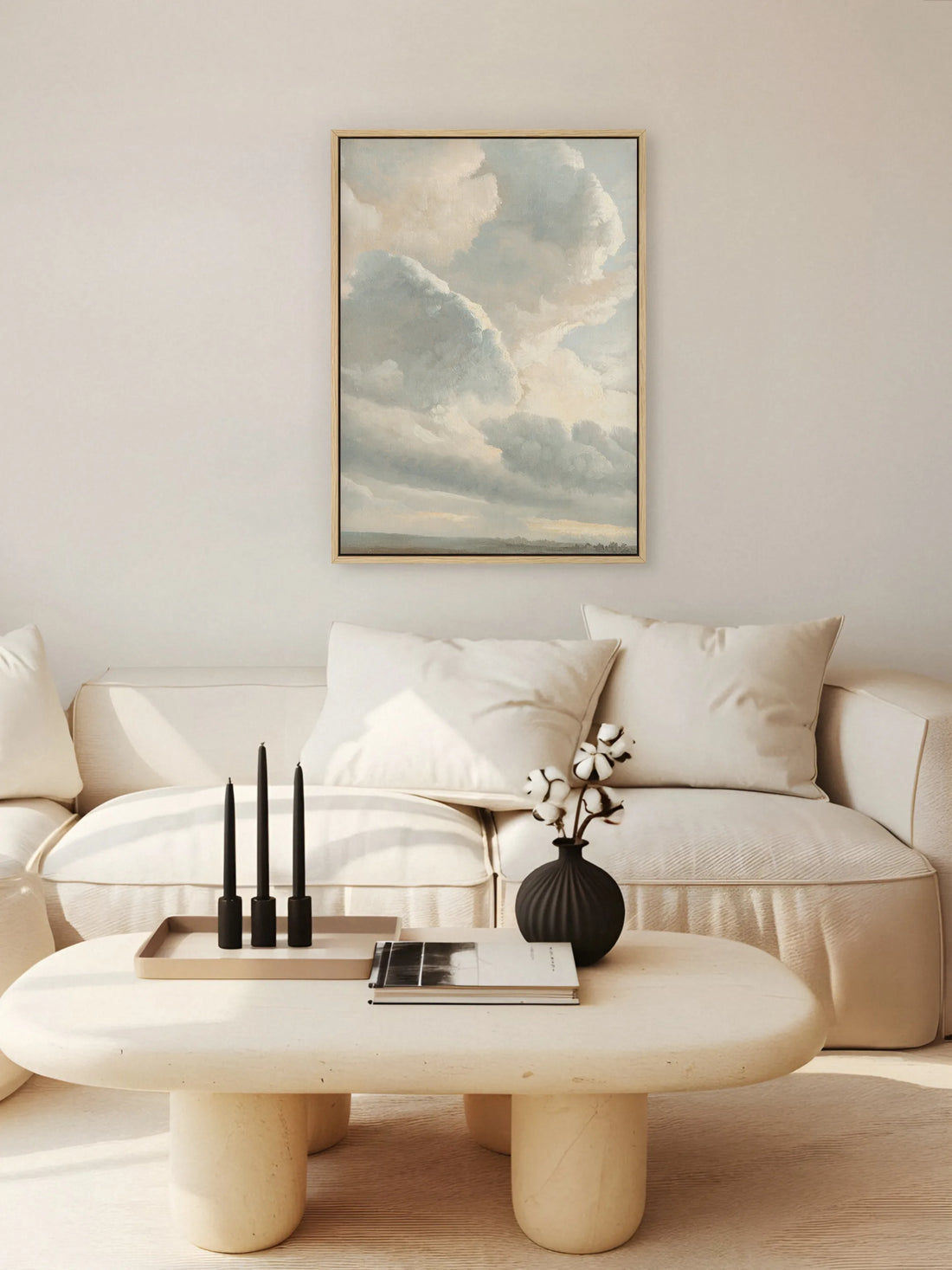 Das Wolken Leinwandbild hängt im Naturholzrahmen über einem cremefarbenen Sofa im Wohnzimmer. Auf dem runden Couchtisch aus hellem Stein stehen drei schwarze Kerzen und eine schwarze Vase mit Baumwollzweigen.