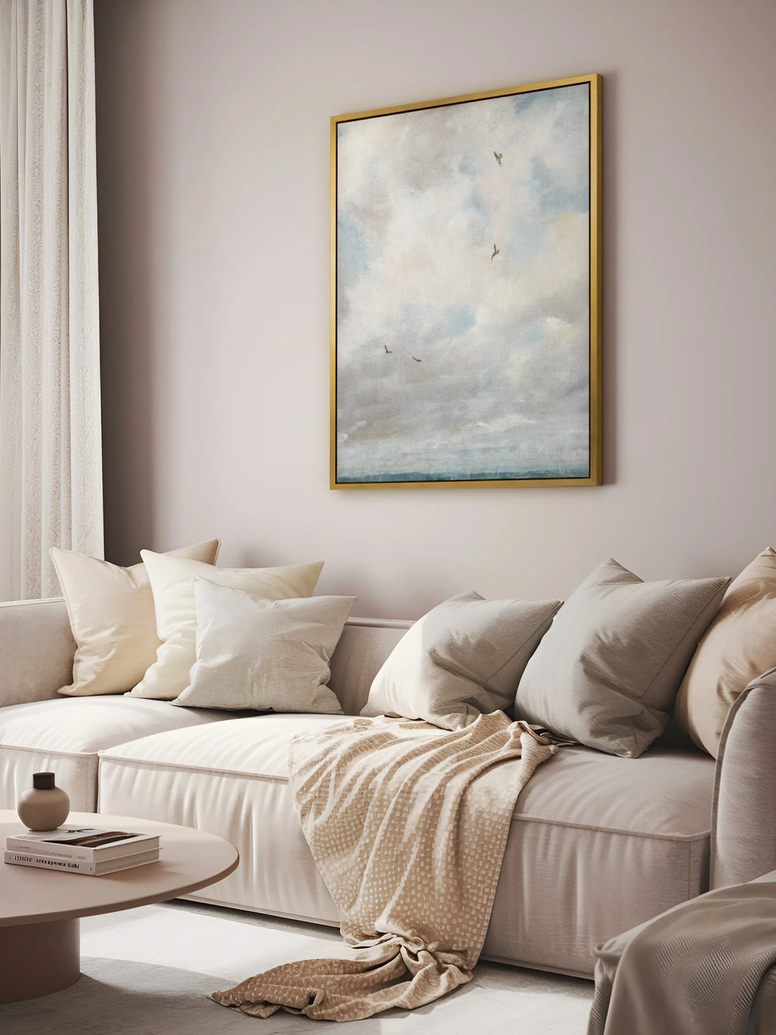 Das Vintage Leinwandbild mit Wolken und Vögeln hängt im goldenen Rahmen an einer greigefarbenen Wand über einem cremefarbenen Sofa. Auf dem Sofa liegen viele Kissen und eine beige Strickdecke, davor steht ein runder Couchtisch.