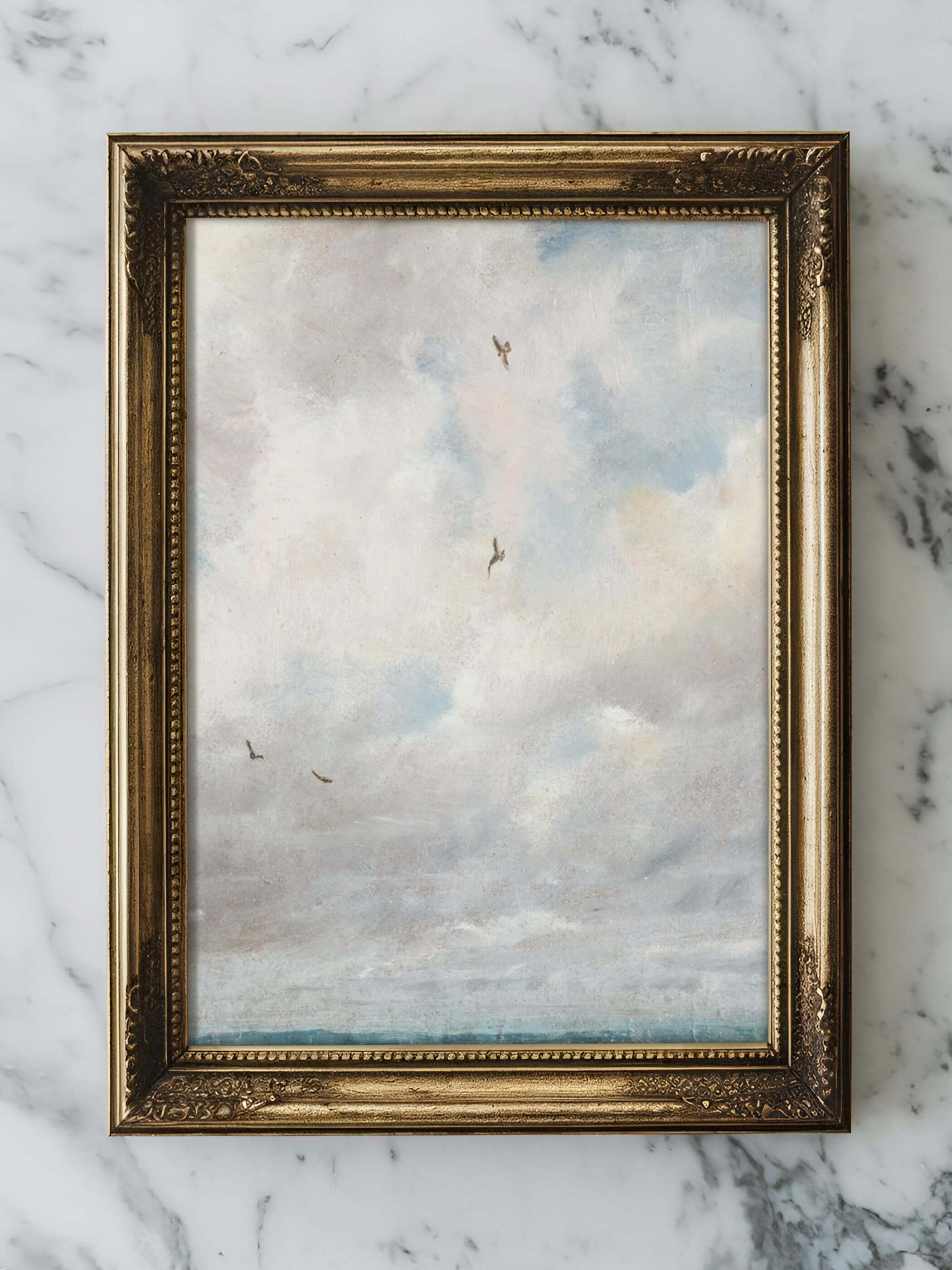 Ein gerahmter Kunstdruck einer Vintage-Wolkenszene im klassischen Goldrahmen auf einem Marmortisch platziert. Weiche, helle Wolken füllen das Bild, das einen ruhigen und friedlichen Eindruck vermittelt.