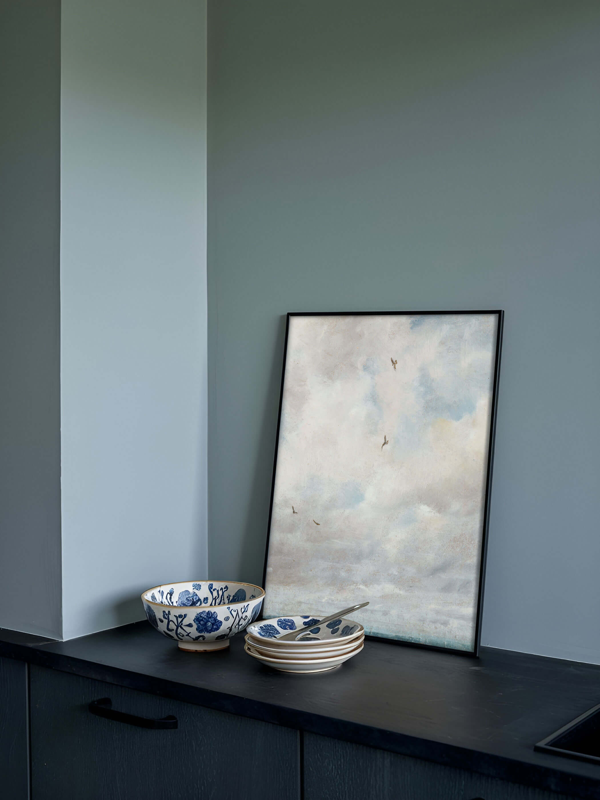 Ein Vintage-Wolkenposter, das auf einer dunklen Küchenablage gegen eine graue Wand lehnt, dekoriert mit blau-weißen Schalen im Vordergrund. Der Kunstdruck bringt Ruhe und Eleganz in die moderne Küche.