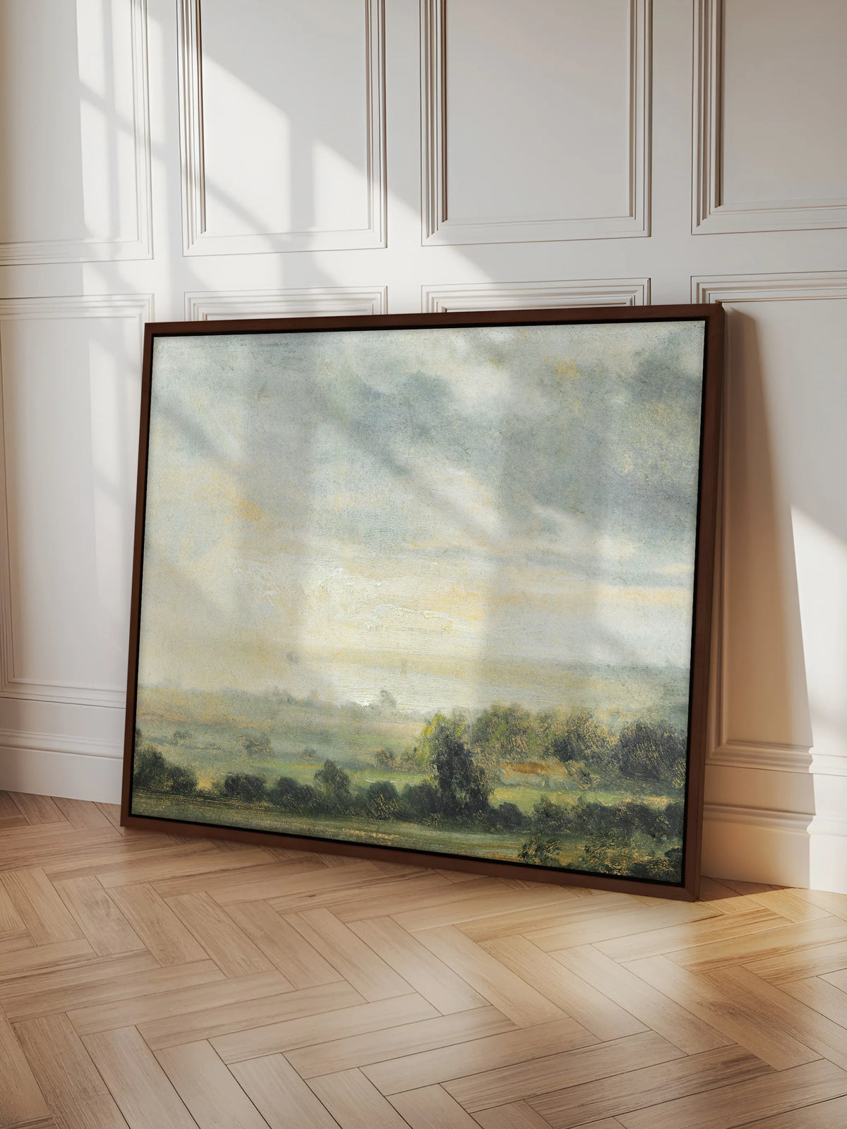 Großes Leinwandbild mit Wolkenhimmel und grünem Tal im braunen Schattenfugenrahmen, angelehnt an eine weiße Kassettenwand. Es steht auf hellem Fischgrätparkett, Sonnenlicht fällt schräg über Bild und Wand.