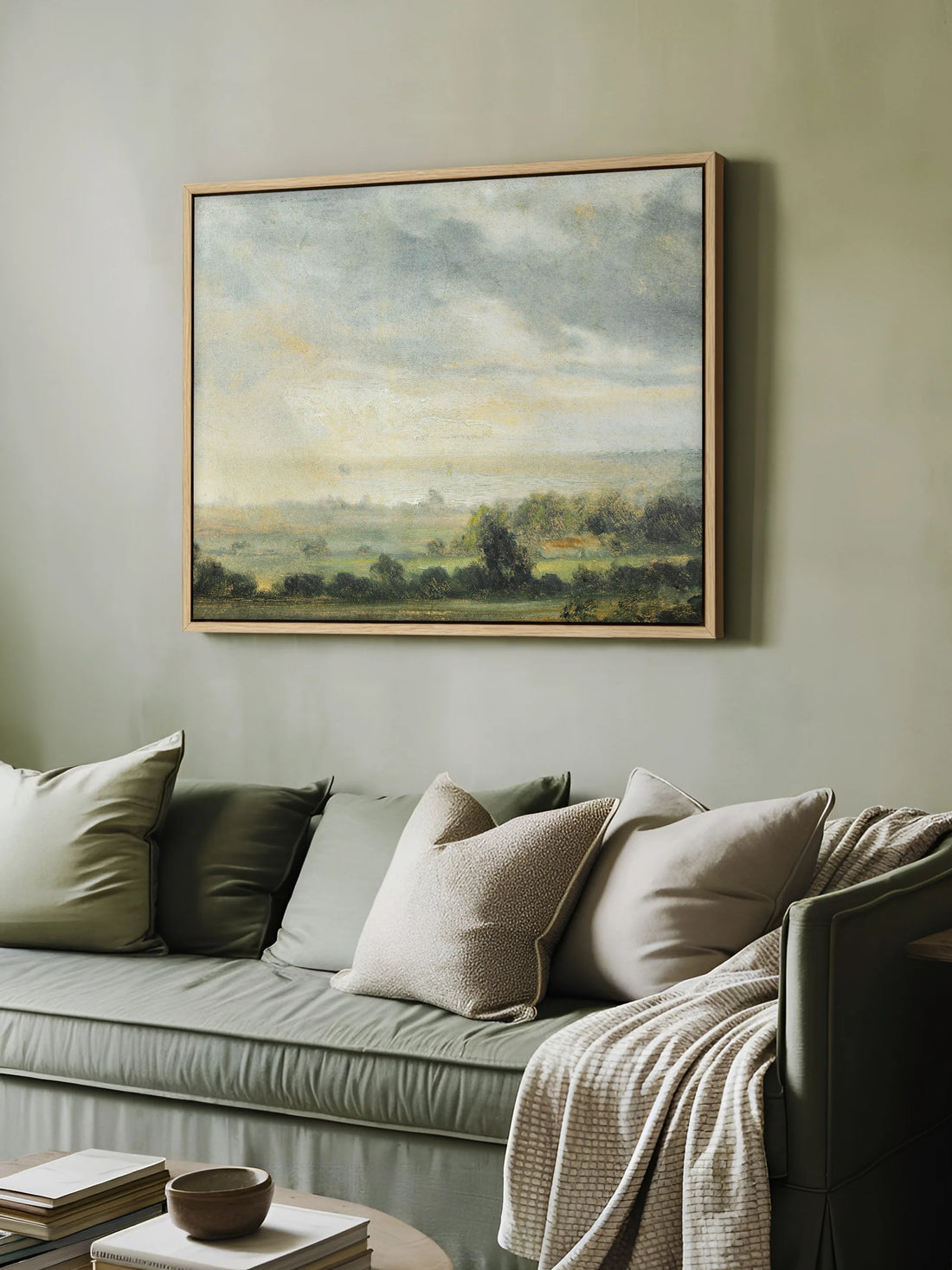 Das Cottagecore Leinwandbild mit weiter Landschaft hängt im Naturholzrahmen an einer salbeigrünen Wand über einem grünen Sofa. Auf dem Sofa liegen Kissen in Grün und Beige und eine cremefarbene Strickdecke.
