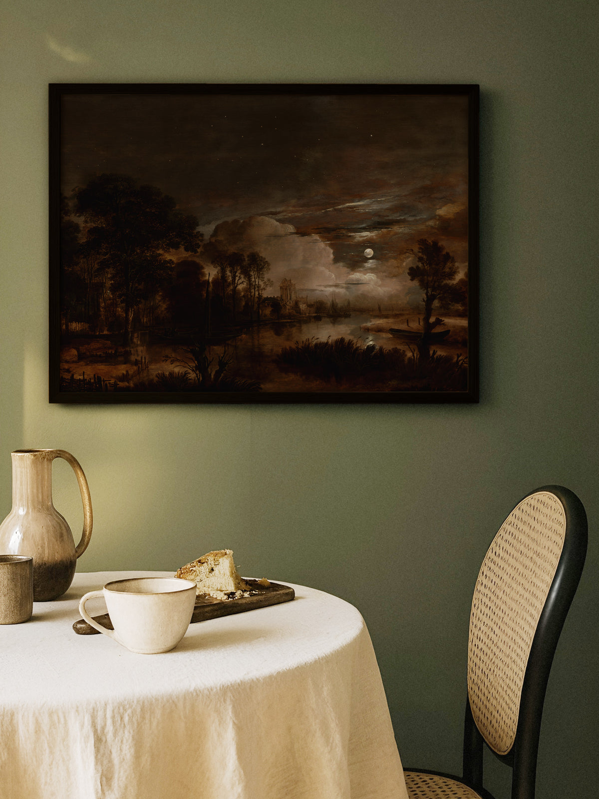 Nachtlandschaft mit Vollmond als Poster im schwarzen Rahmen an einer salbeigrünen Wand über einem runden Tisch mit Leinentuch, Keramikkrug und Kaffeetasse. Die tiefen Brauntöne passen zum moody Esszimmer.