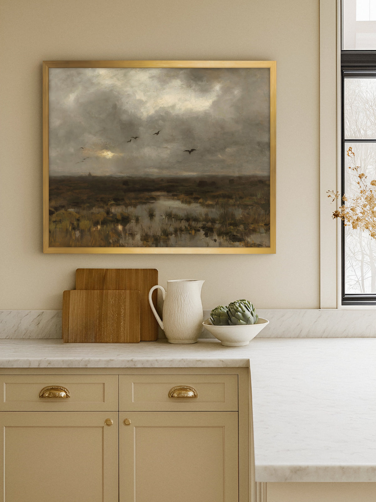 Moorlandschaft mit Vögeln als Kunstdruck im goldenen Rahmen über einer Marmorarbeitsplatte mit Holzbrettern, weißem Krug und Artischocken in einer beigen Küche.
