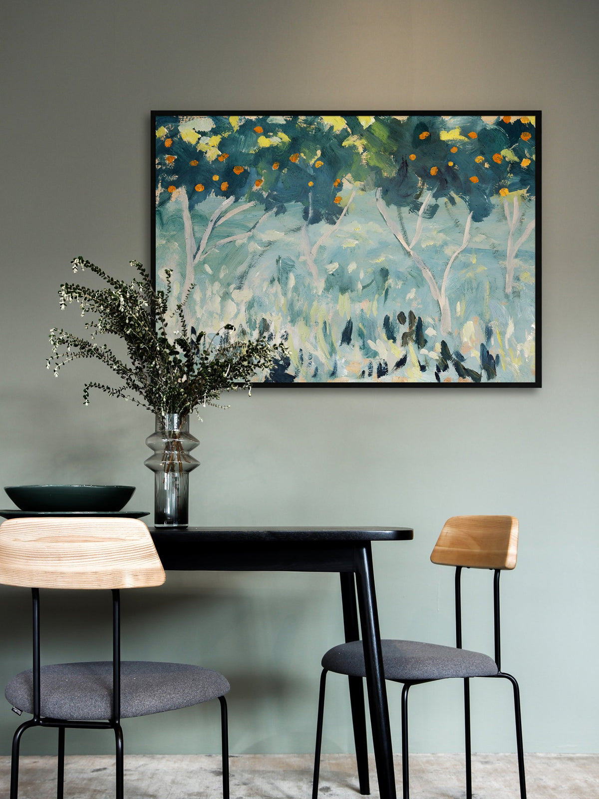 Wandbild mit Orangenbäumen im schwarzen Rahmen an einer salbeigrünen Wand über einem schwarzen Esstisch mit Glasvase und grünen Zweigen, davor zwei Stühle mit Holzlehnen. Das Petrol des Motivs passt zum gedeckten Grün des Esszimmers.