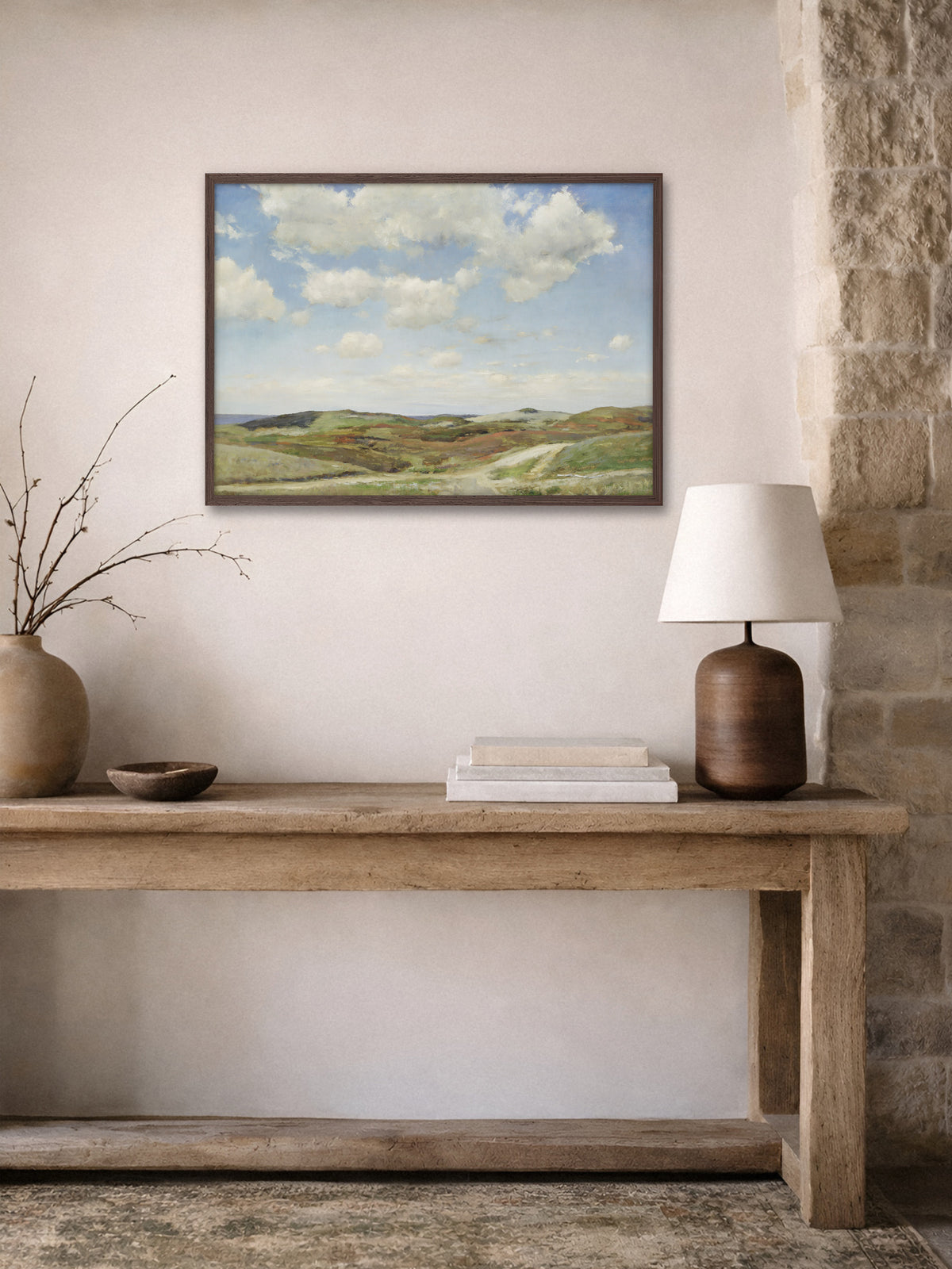 Poster einer Dünenlandschaft im dunklen Holzrahmen über einer rustikalen Holzkonsole mit Tonvase, Holzschale, Büchern und Tischlampe neben einer Natursteinwand. Das Vintage Motiv bringt Himmel und Weite in den Flur im Wabi-Sabi Stil.