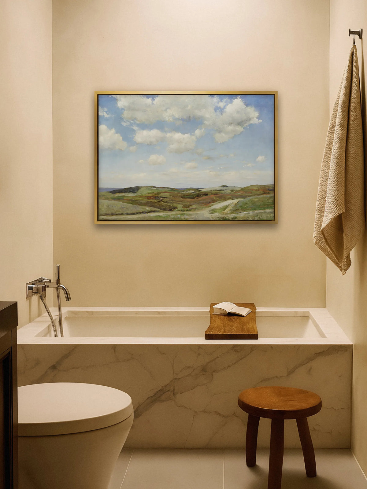 Küstenlandschaft als Leinwandbild im goldenen Rahmen über einer Badewanne mit Marmorverkleidung. Auf einem Holzbrett über der Wanne liegt ein offenes Buch, davor steht ein runder Holzhocker, rechts hängt ein Leinenhandtuch.