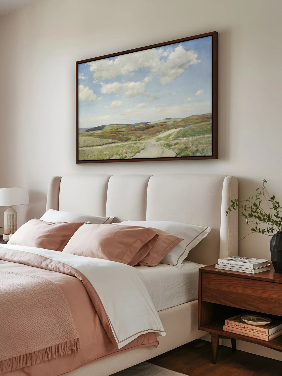 Großes Leinwandbild mit Hügeln und Wolkenhimmel im braunen Rahmen über einem Polsterbett in Creme mit rosé und weißer Bettwäsche. Daneben steht ein Nachttisch aus Nussbaum mit Büchern und einer schwarzen Vase mit grünen Zweigen.
