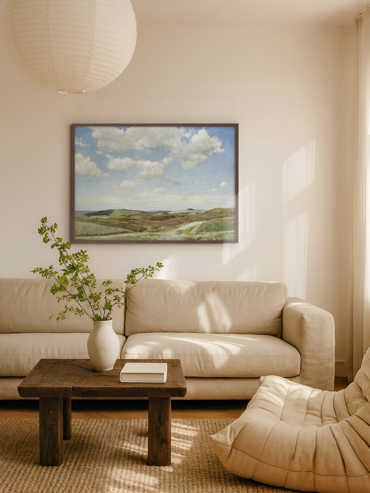 Kunstdruck mit Dünenlandschaft im dunklen Holzrahmen über einem beigen Sofa, davor ein rustikaler Holztisch mit weißer Vase und grünen Zweigen, darüber eine Papierleuchte. Die weite Küstenlandschaft passt zum hellen Wohnzimmer im Japandi Stil.