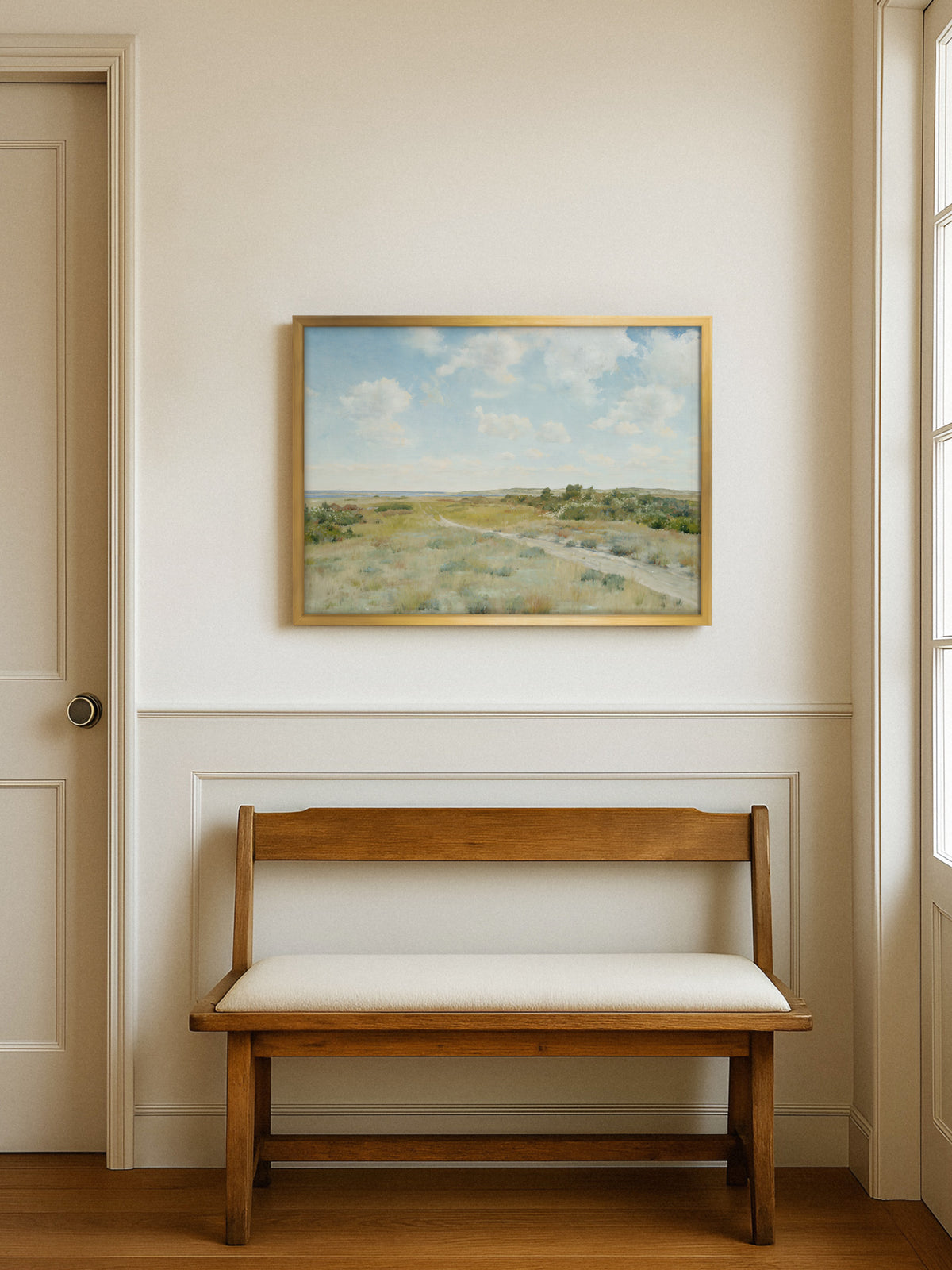 Poster einer Heidelandschaft im goldenen Rahmen an einer cremeweißen Wand mit Wandvertäfelung über einer Holzbank mit hellem Sitzpolster, daneben eine Tür und ein Fenster. Die Vintage Landschaft passt zum klassisch eingerichteten Flur.