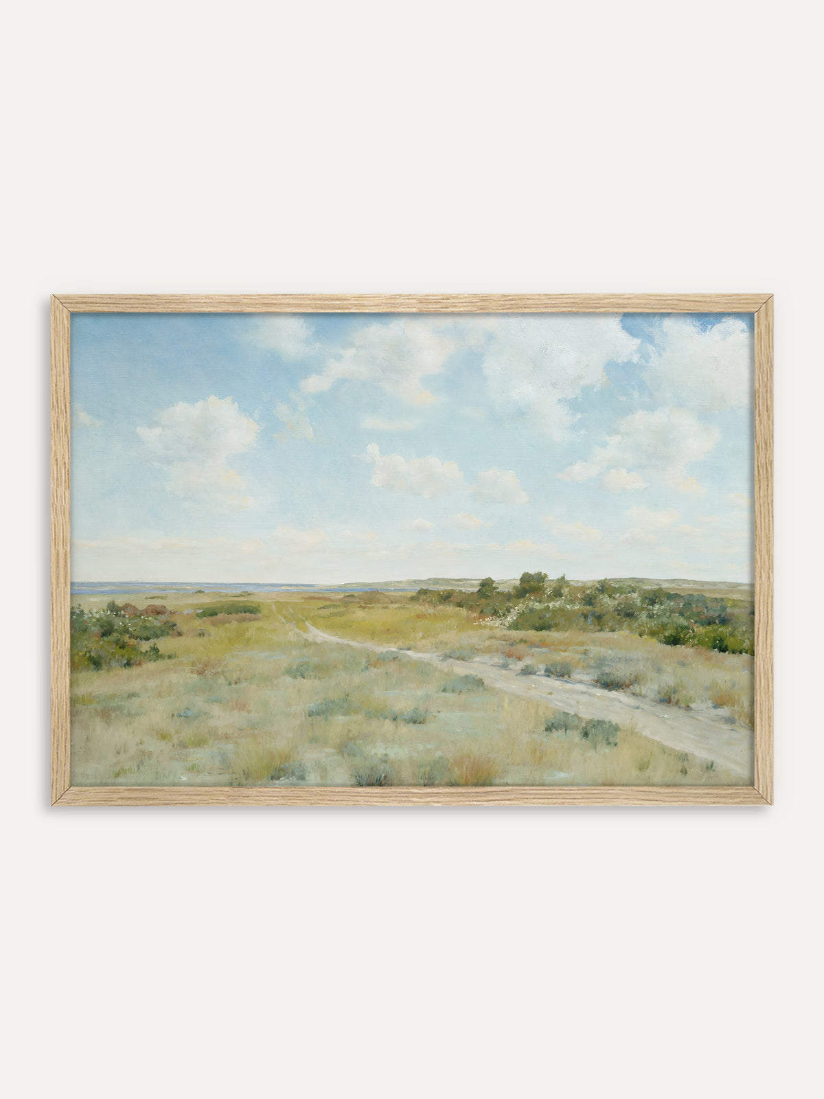 Kunstdruck, der eine Heidelandschaft mit einem sandigen Weg durch salbeigrünes Gras und blühende Büsche zeigt, am Horizont blaues Wasser unter weißen Wolken. Im Rahmen aus hellem Holz bleibt das Motiv leicht und passt zu einem Landhaus am Meer. #Rahmen_Helles Holz