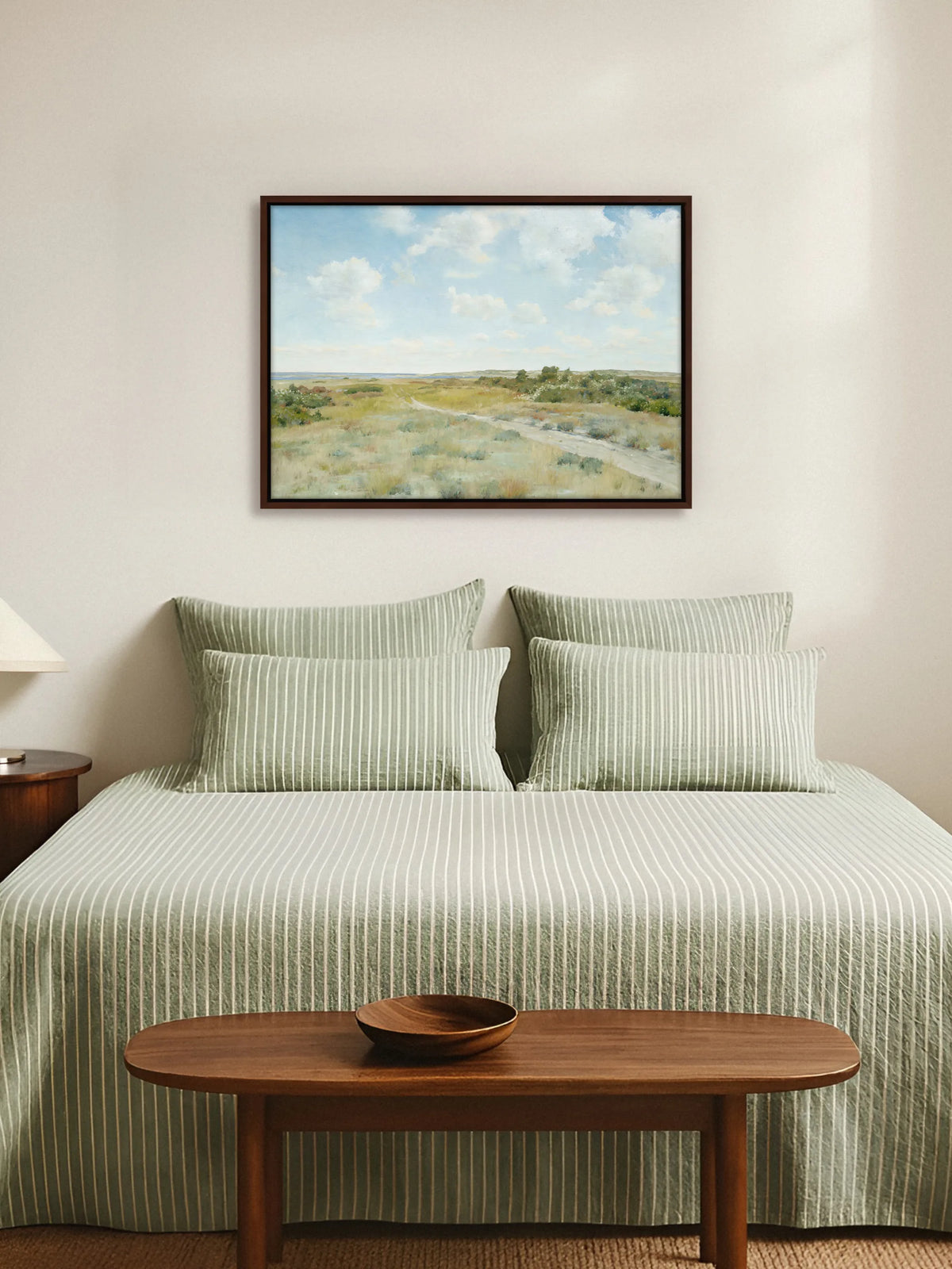 Leinwandbild mit Weg durch die Heide im braunen Rahmen über einem Bett mit salbeigrün gestreifter Leinenbettwäsche. Am Fußende steht eine ovale Holzbank mit einer Holzschale, die Wand ist cremeweiß.