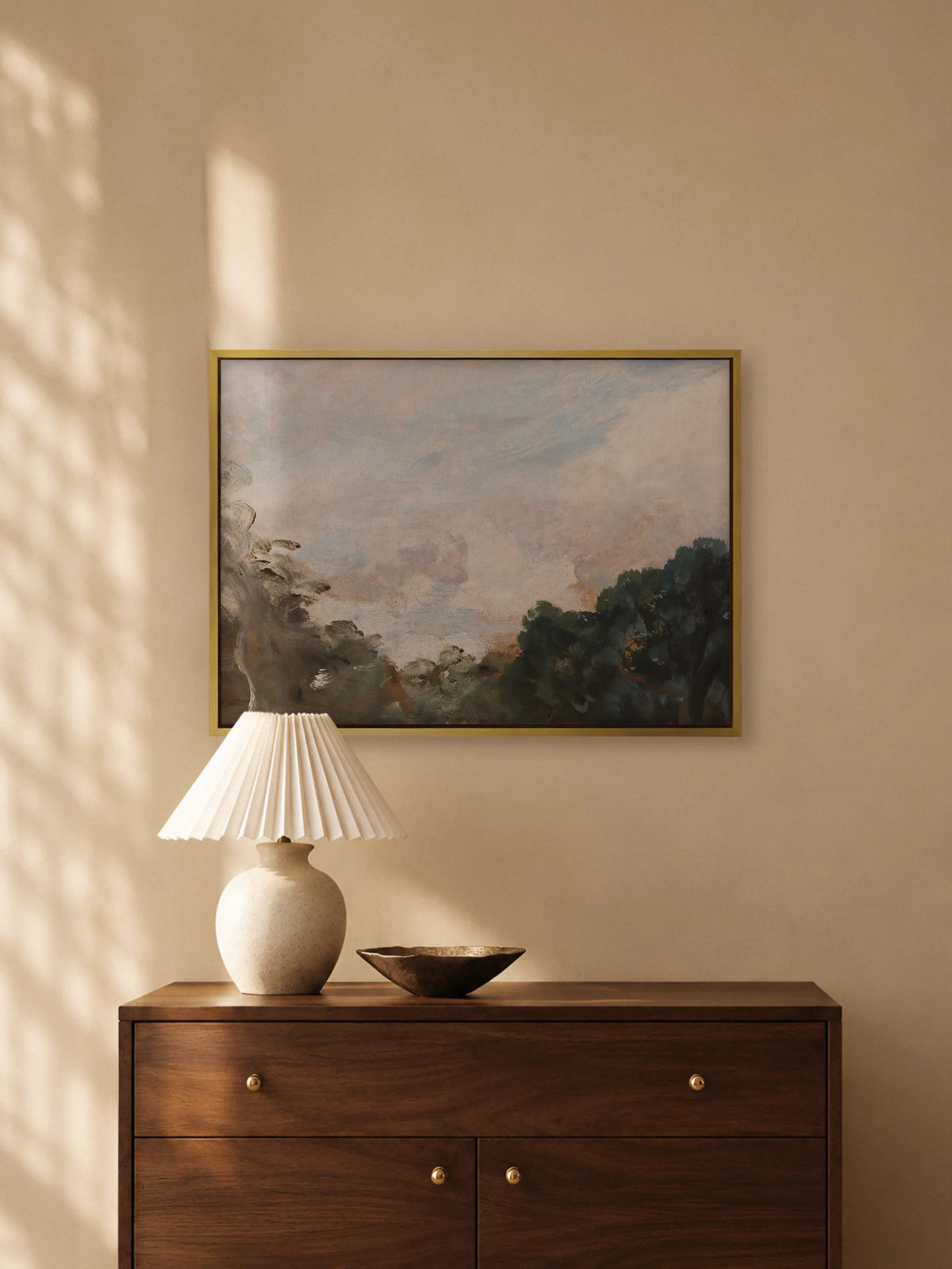 Wolkenbild auf Leinwand im goldenen Rahmen über einer dunklen Holzkommode mit Messingknöpfen. Auf der Kommode stehen eine Keramiklampe mit weißem Plisseeschirm und eine flache Bronzeschale, warmes Licht fällt auf die Wand.