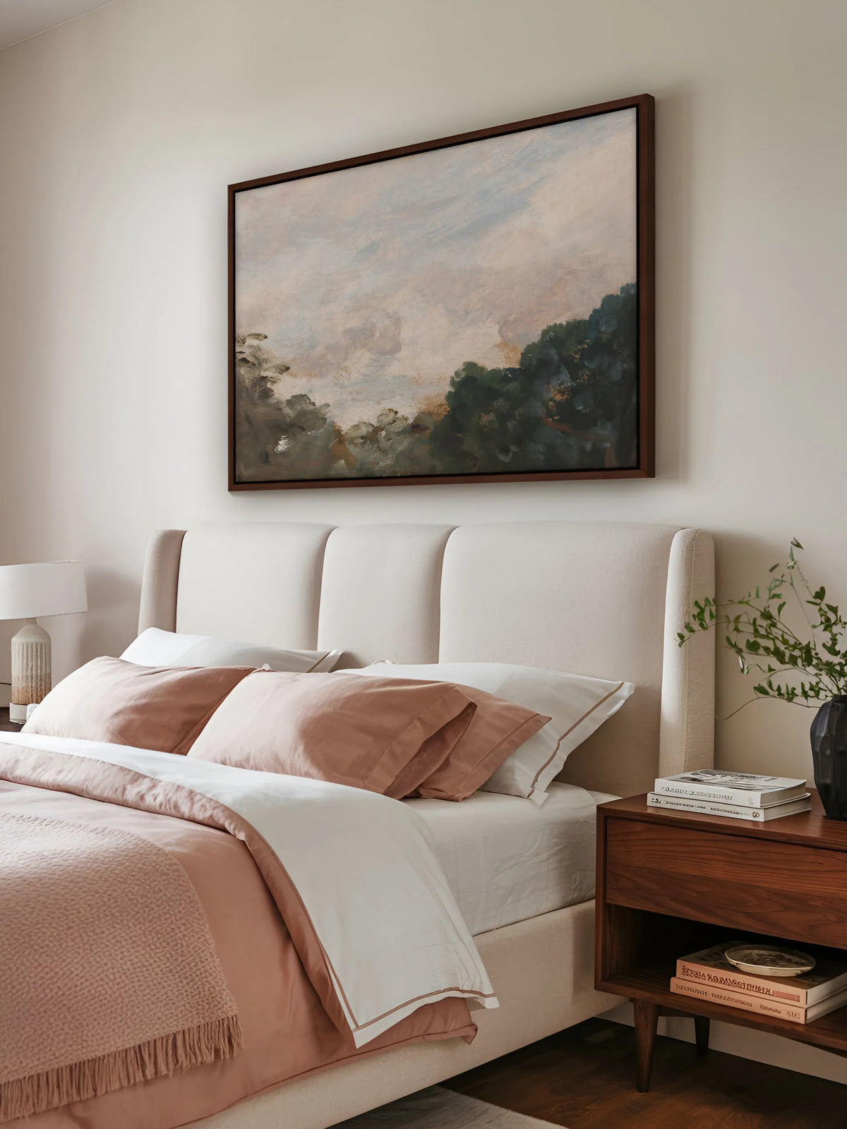 Großes Leinwandbild mit rosigem Wolkenhimmel im braunen Rahmen über einem Polsterbett in Creme mit rosé und weißer Bettwäsche. Daneben steht ein Nachttisch aus Nussbaum mit Büchern und einer schwarzen Vase mit grünen Zweigen.
