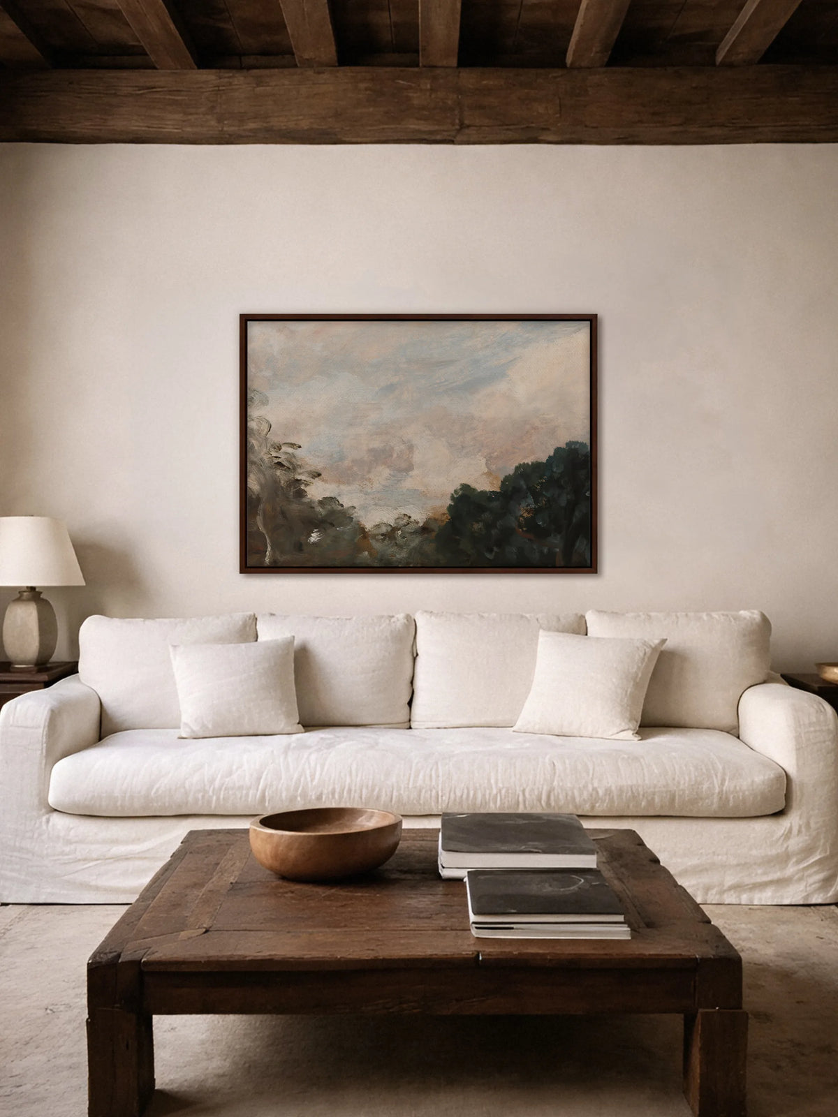 Das Vintage Leinwandbild mit Wolken über Bäumen hängt im braunen Rahmen über einem weißen Leinensofa unter dunklen Holzbalken. Davor steht ein rustikaler Couchtisch aus altem Holz mit einer Holzschale und Büchern.
