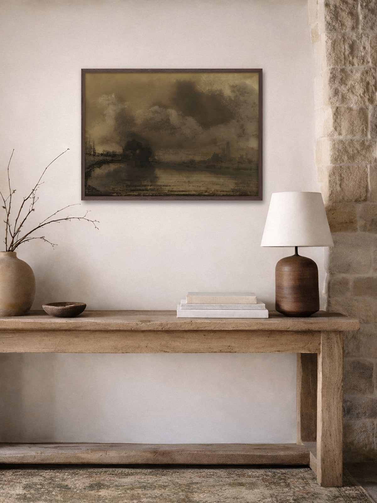 Wandbild einer Uferlandschaft in Sepia im dunklen Holzrahmen über einer rustikalen Holzkonsole mit Tonvase, Holzschale, Büchern und Tischlampe neben einer Natursteinwand. Die Erdtöne passen zum Flur im Wabi Sabi Stil.