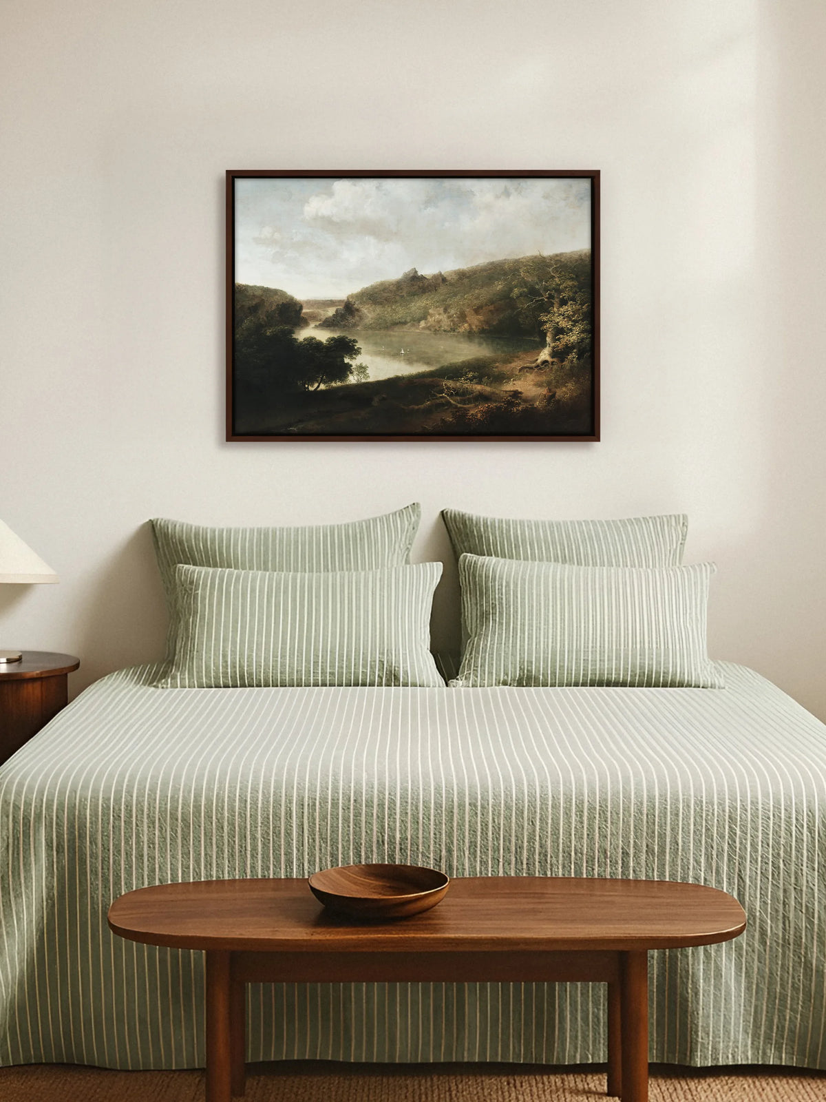 Seelandschaft als Leinwandbild im braunen Rahmen über einem Bett mit salbeigrün gestreifter Leinenbettwäsche. Am Fußende steht eine ovale Holzbank mit einer Holzschale, die Wand ist cremeweiß.