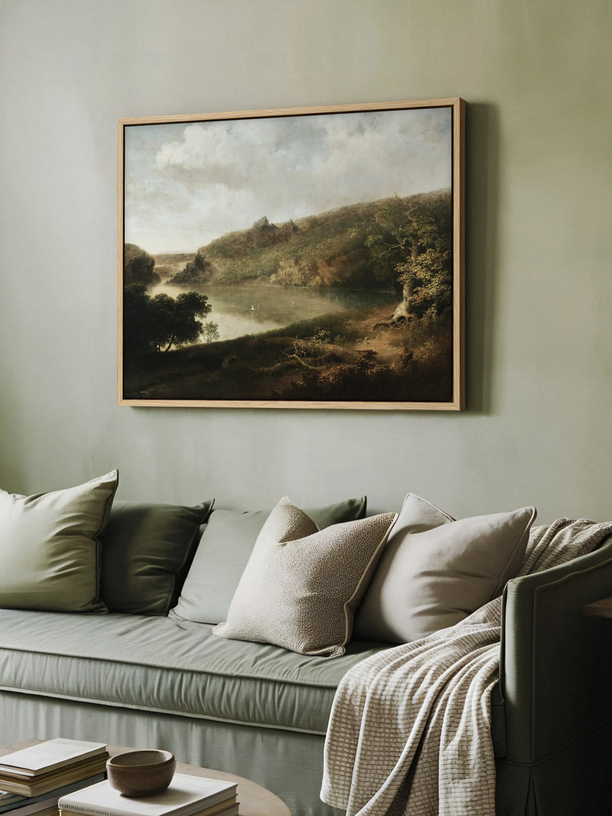 Das Vintage Leinwandbild mit Seelandschaft hängt im Naturholzrahmen an einer salbeigrünen Wand über einem grünen Sofa. Auf dem Sofa liegen Kissen in Grün und Beige und eine cremefarbene Strickdecke, davor Bücher und eine Schale.
