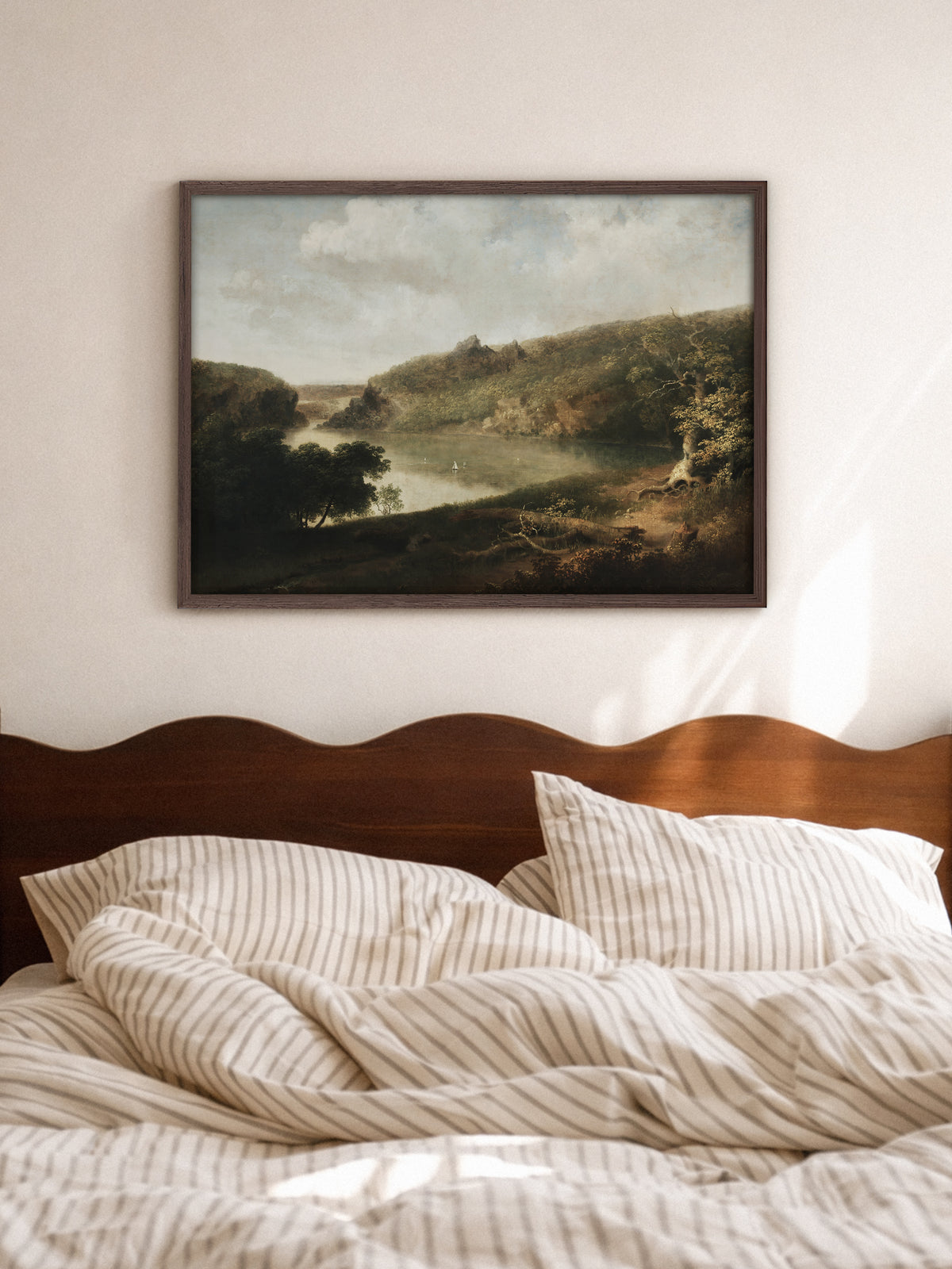 Wandbild einer Seelandschaft im dunklen Holzrahmen über einem Bett mit geschwungenem Kopfteil aus Holz und gestreifter Bettwäsche. Das ruhige Vintage Motiv passt zum hellen Schlafzimmer.