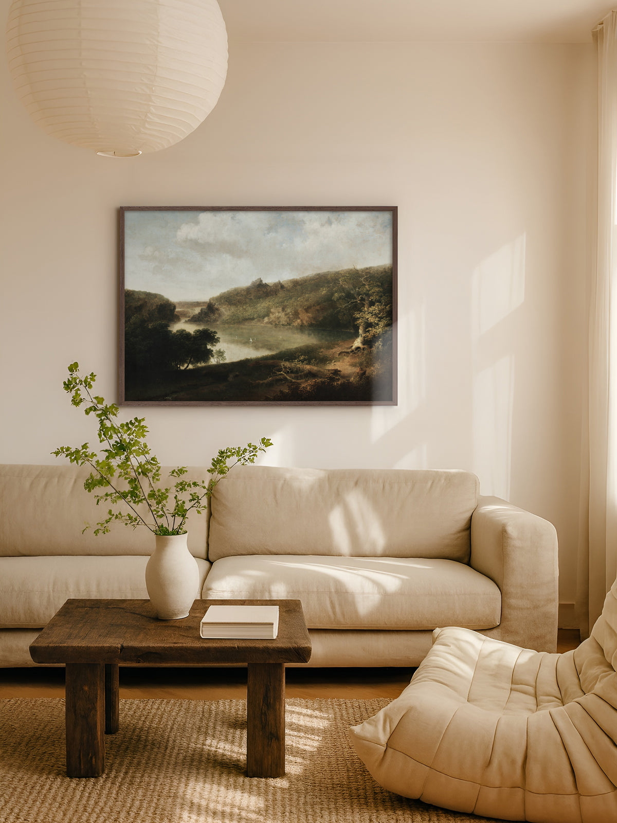 Kunstdruck einer Seelandschaft im dunklen Holzrahmen über einem beigen Sofa, davor ein rustikaler Holztisch mit weißer Vase und grünen Zweigen, darüber eine Papierleuchte. Die Erdtöne des Motivs passen zum hellen Wohnzimmer im Japandi Stil.