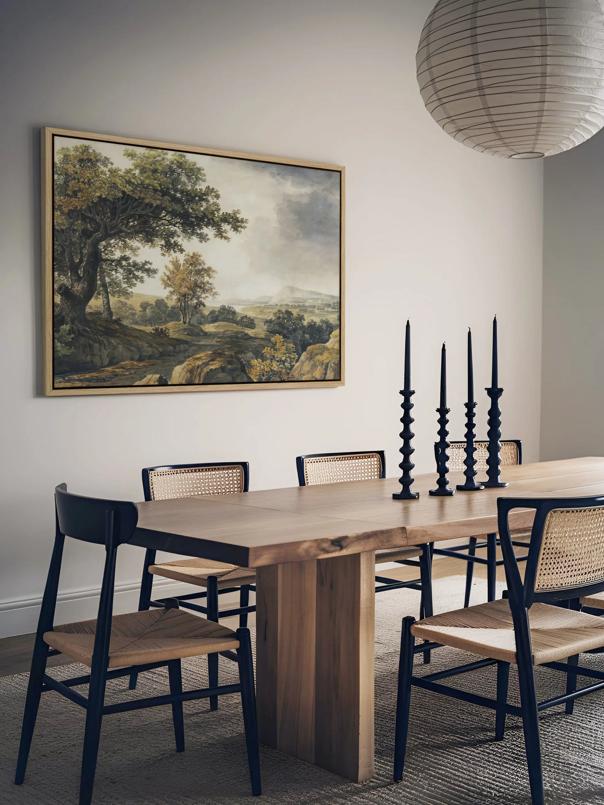Das Vintage Leinwandbild mit Waldlandschaft hängt im Naturholzrahmen an einer hellgrauen Wand im Esszimmer. Davor stehen ein massiver Holztisch mit schwarzen Wiener Geflechtstühlen und vier schwarze Kerzenleuchter, darüber eine Papierlampe.