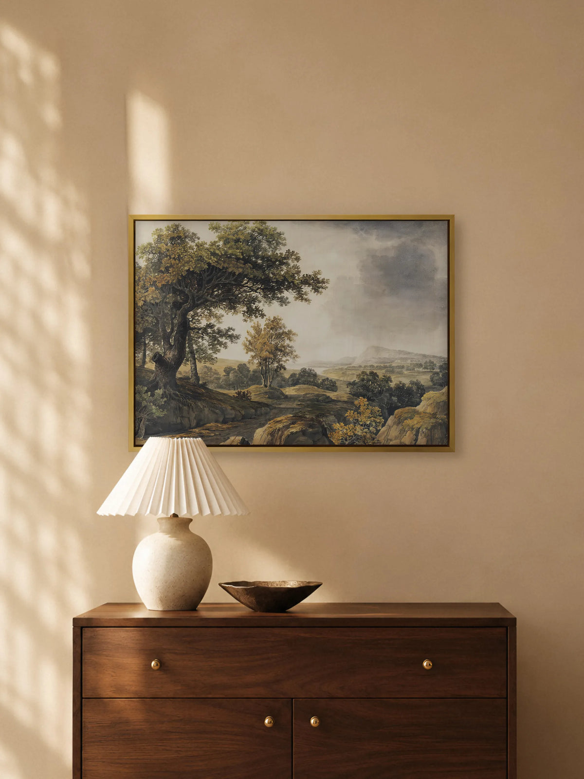 Waldlandschaft auf Leinwand im goldenen Rahmen über einer dunklen Holzkommode mit Messingknöpfen. Auf der Kommode stehen eine Keramiklampe mit weißem Plisseeschirm und eine flache Bronzeschale, warmes Licht fällt auf die Wand.